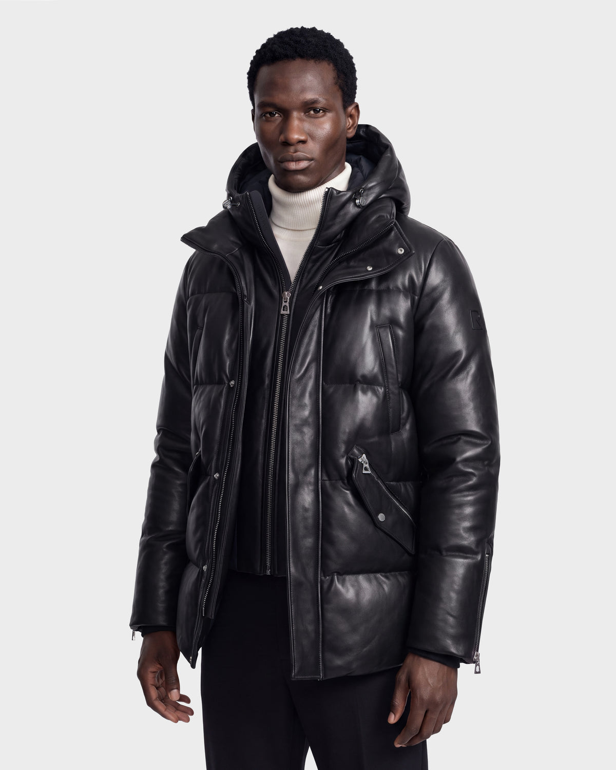 A man wearing the Rudsak APOLLO X - 6125696 Black leather puffer jacket over a white turtleneck sweater stands against a plain light gray background, looking at the camera with a neutral expression.