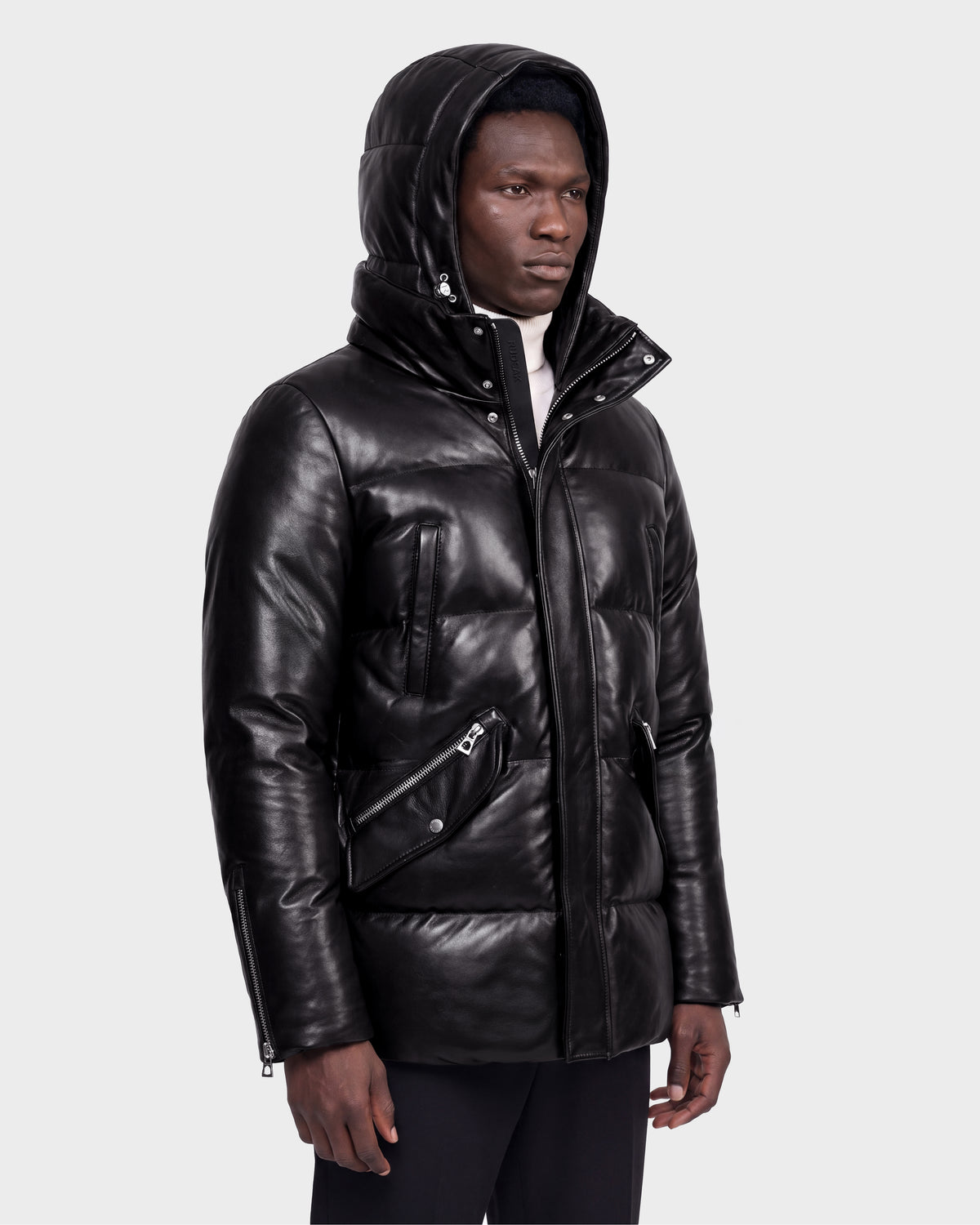 A man stands against a plain light gray background wearing the Rudsak APOLLO X - 6125696 Black leather hooded puffer jacket, featuring zipper and snap details.