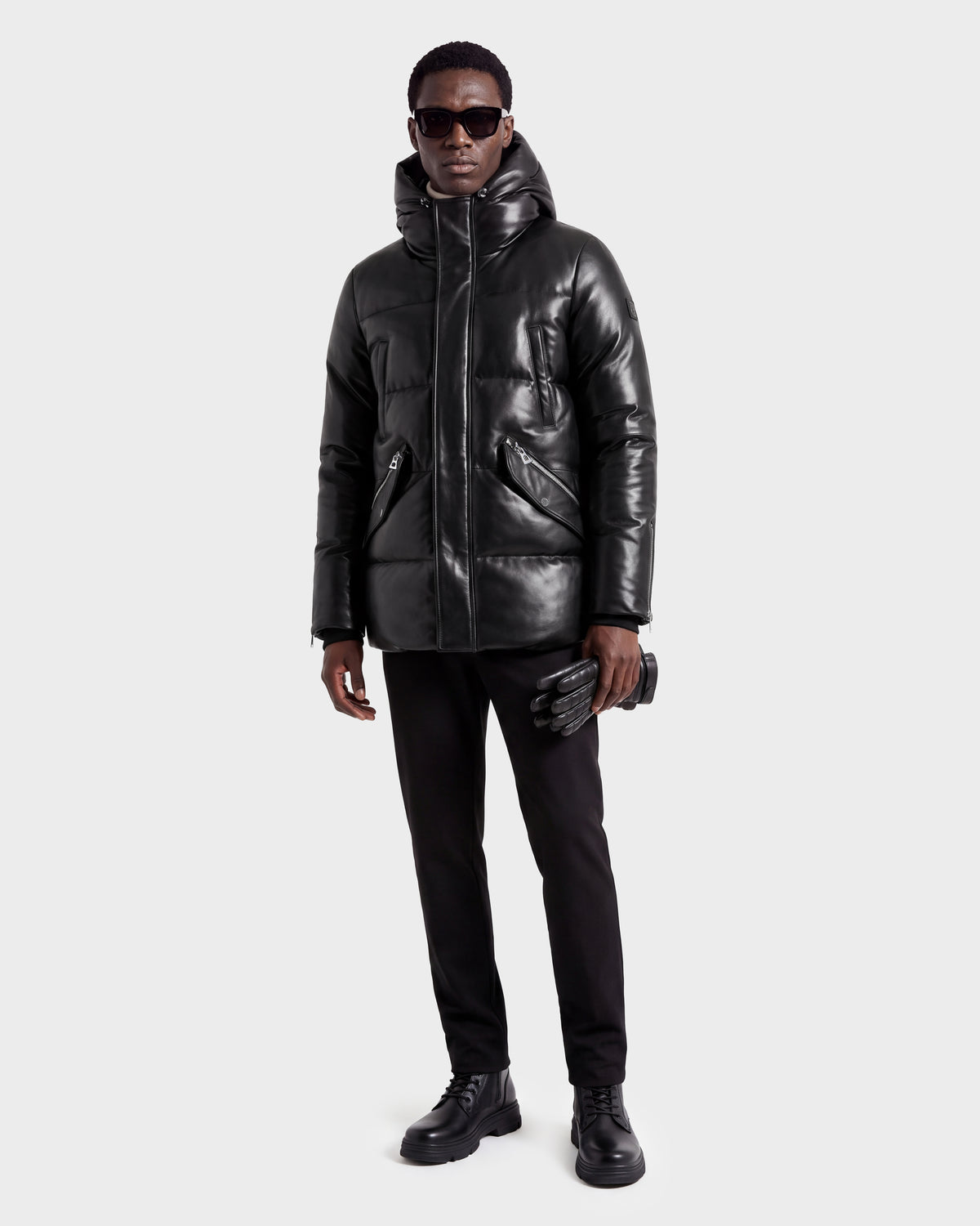 A man wearing the Rudsak APOLLO X - 6125696 Black hooded leather jacket, paired with black pants, black shoes, dark sunglasses, and holding black gloves stands against a plain light background.