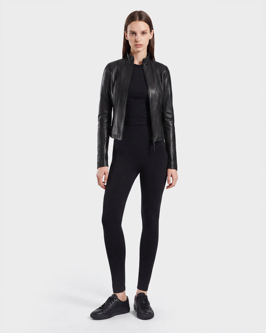 A person stands facing forward against a plain white background, dressed in the Rudsak ABIGAIL - 8124560 Black women’s leather jacket, styled with a black top, black leggings, and black sneakers.