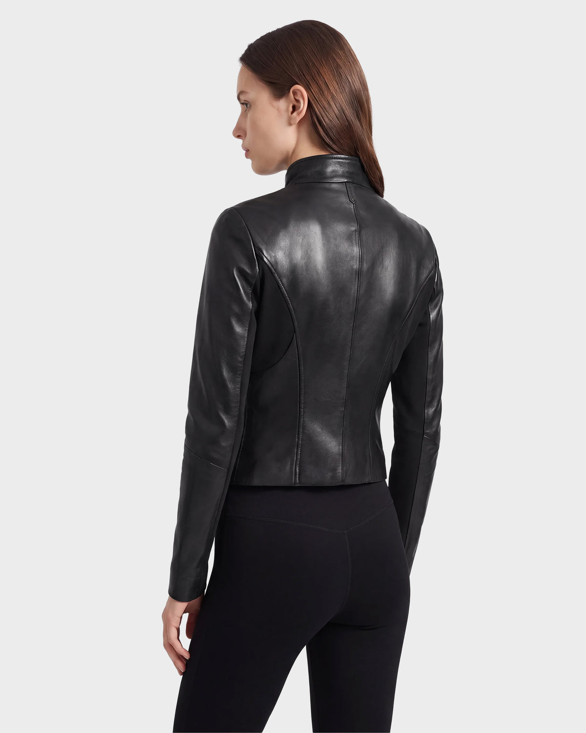 A woman with straight brown hair is wearing the Rudsak ABIGAIL - 8124560 Black women's leather jacket and black pants, standing with her back partially turned against a plain light gray background.