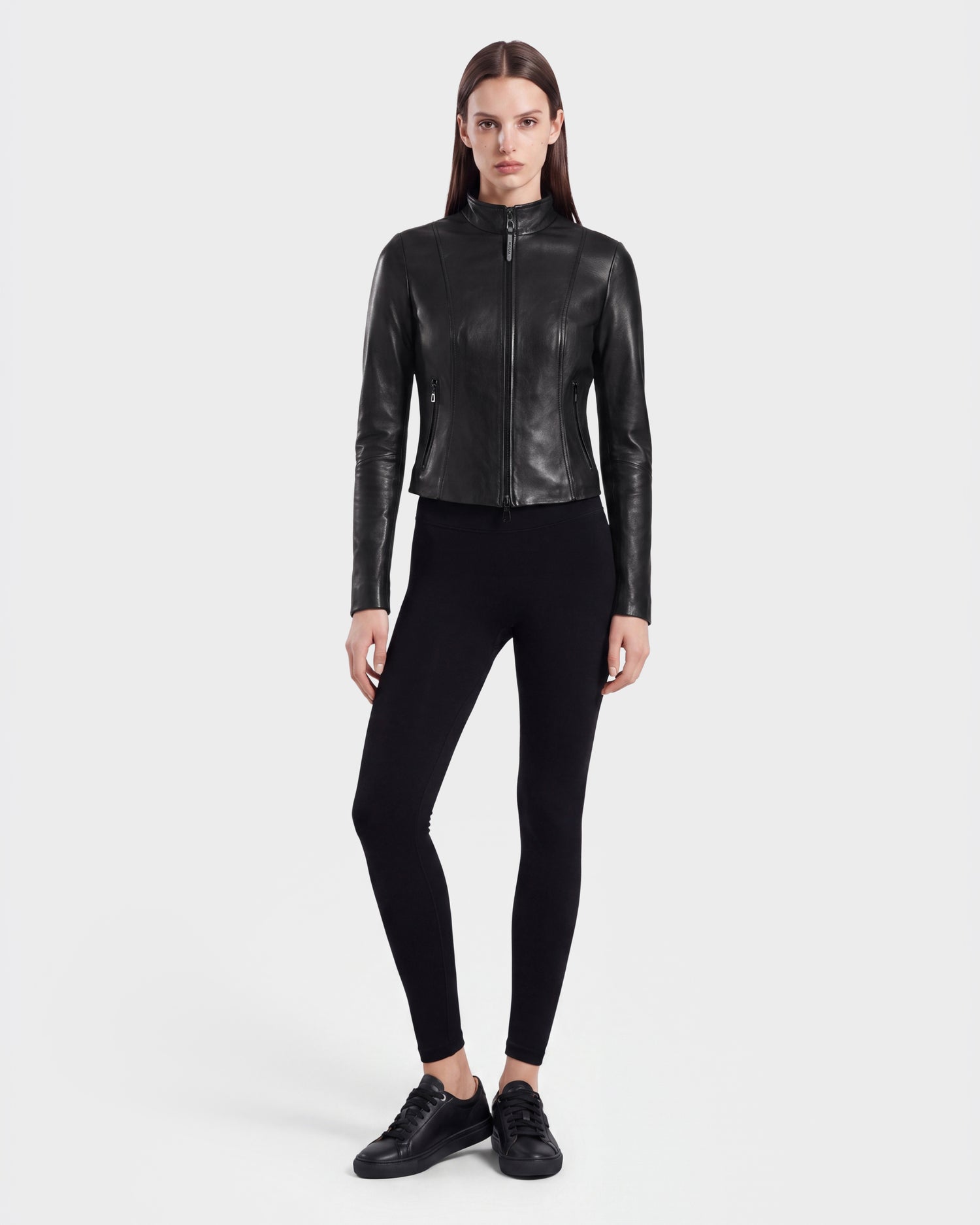 A person stands facing forward, wearing the Rudsak ABIGAIL - 8124560 Black women's leather jacket styled as a sleek racer jacket, paired with black leggings and black sneakers against a plain white background.