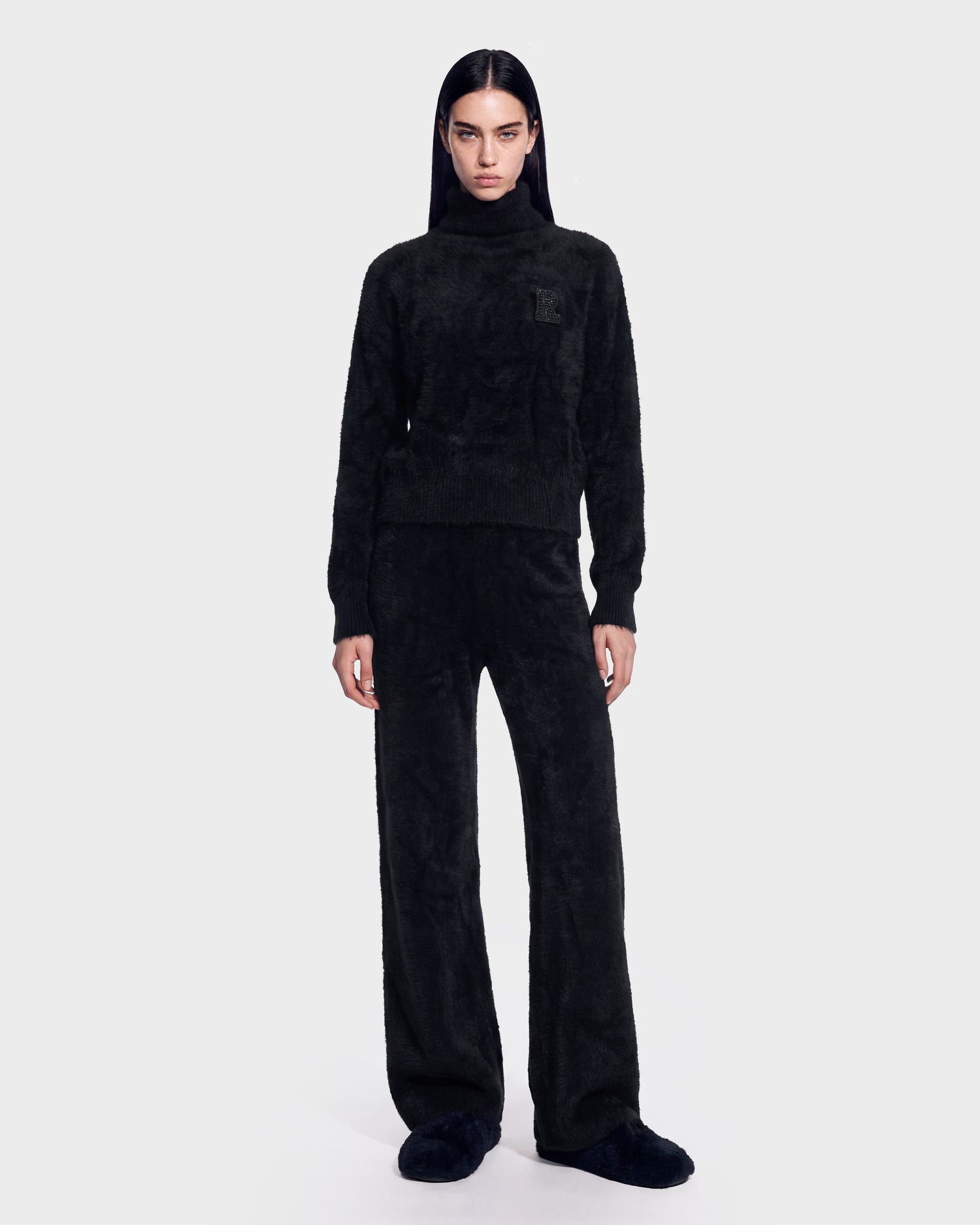 A person stands facing forward, wearing the Rudsak ALISYA - 8526505 Black fuzzy turtleneck sweater with matching wide-leg pants and slippers. The background is plain and light-colored.