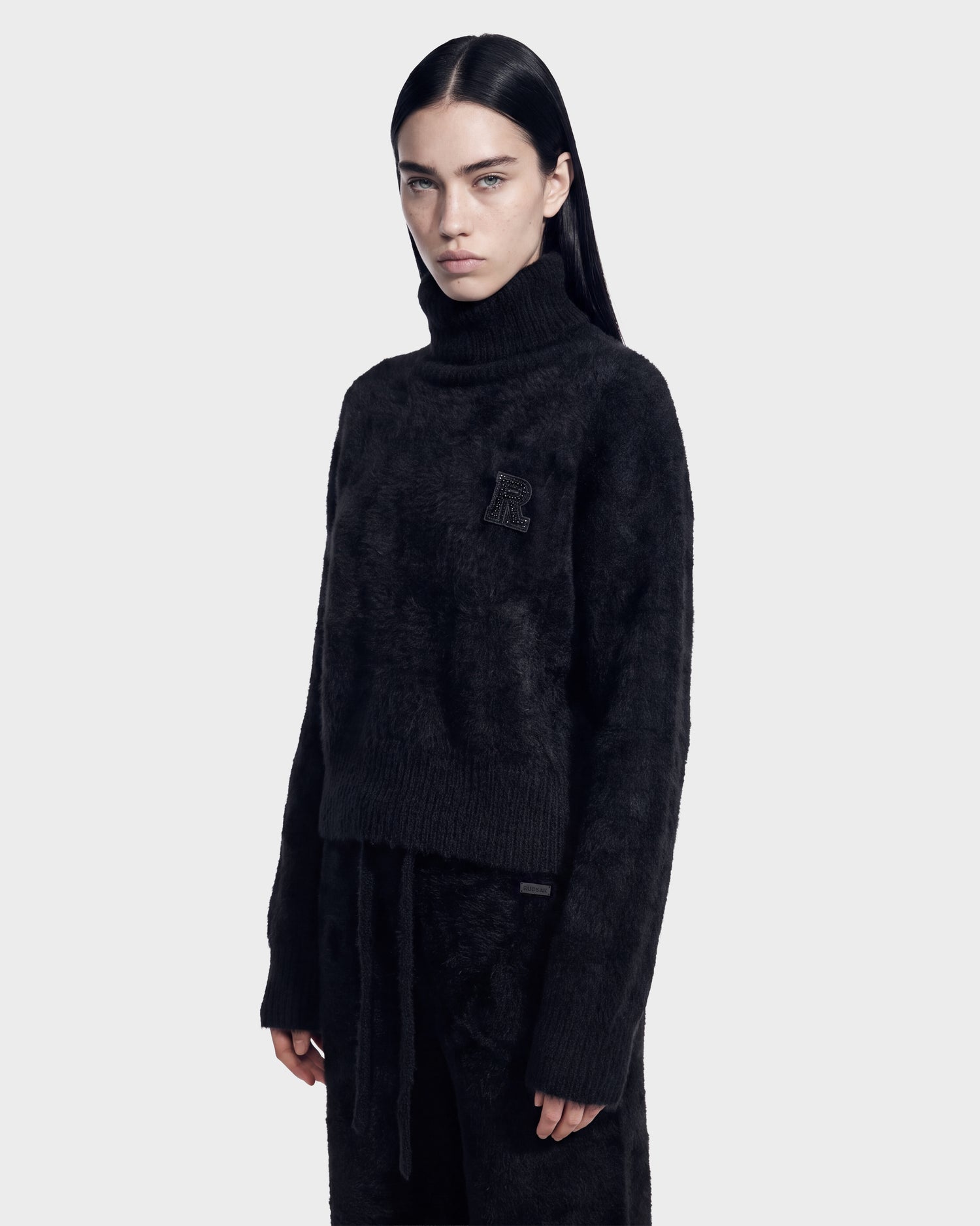 A woman with long dark hair wears the ALISYA - 8526505 Black turtleneck sweater and matching pants by Rudsak. She stands against a plain light gray background, looking slightly to the side with a neutral expression.