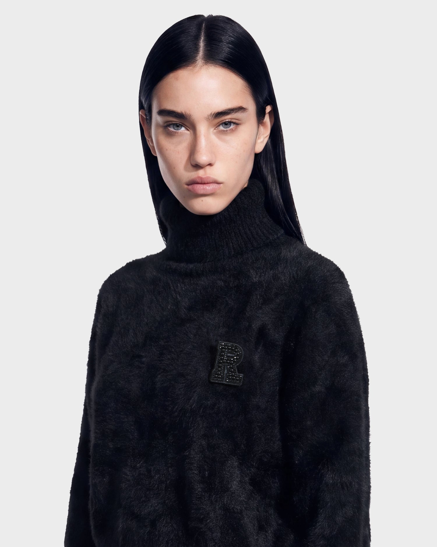 A person with long dark hair is wearing the Rudsak ALISYA - 8526505 Black fuzzy turtleneck sweater, featuring a black letter R patch on the left side of the chest, standing against a plain light gray background.