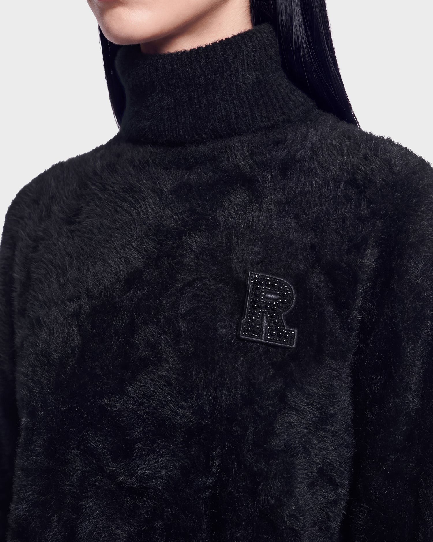 A person wearing the ALISYA - 8526505 Black fuzzy turtleneck sweater by Rudsak, featuring a textured “R” patch on the chest. The person’s face is not visible.