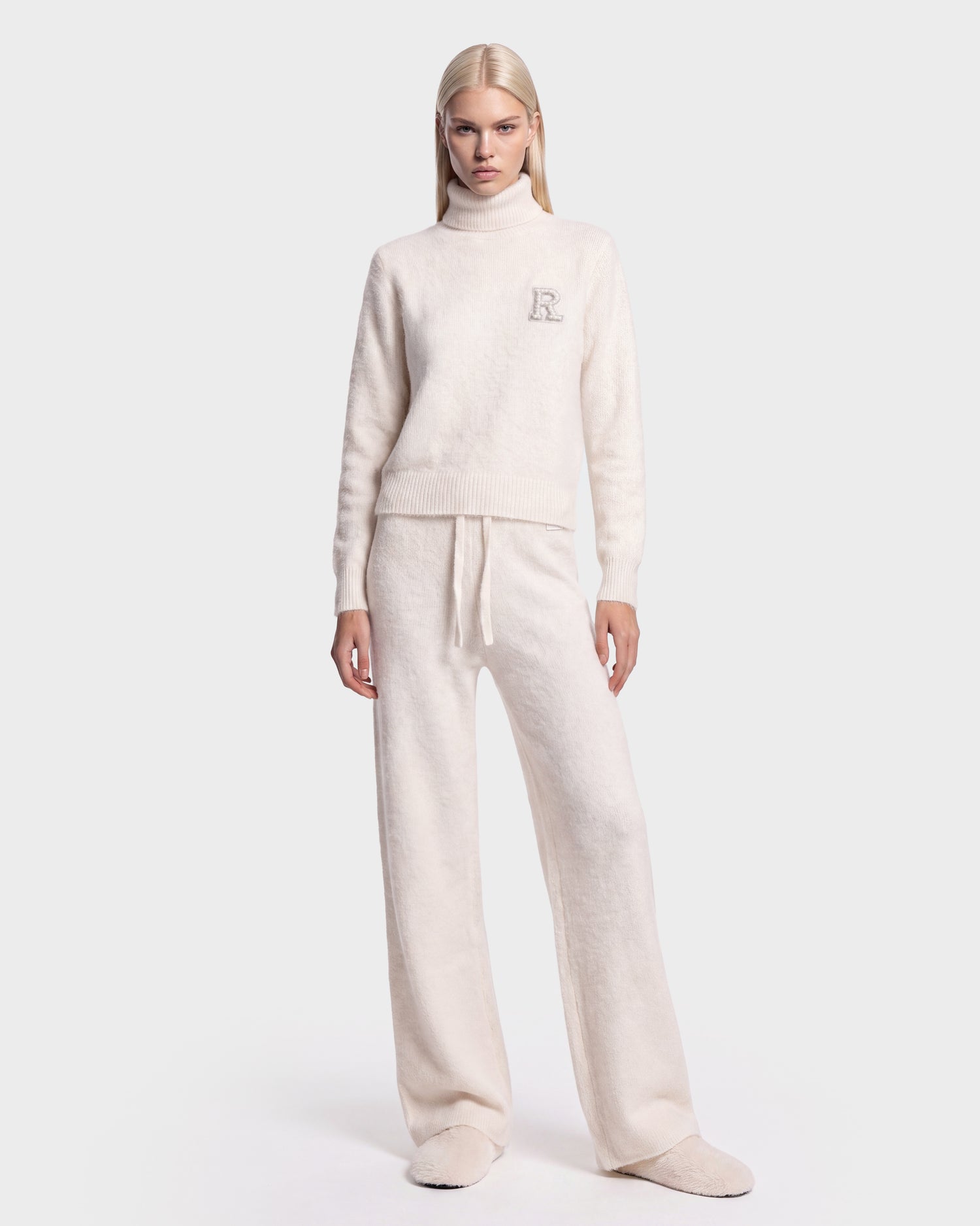 A person stands facing forward against a plain white background, wearing the ALISYA - 8526505 Iceland set by Rudsak: a cozy cream-colored high-neck sweater featuring a small “R” on the chest, paired with matching wide-leg pants.