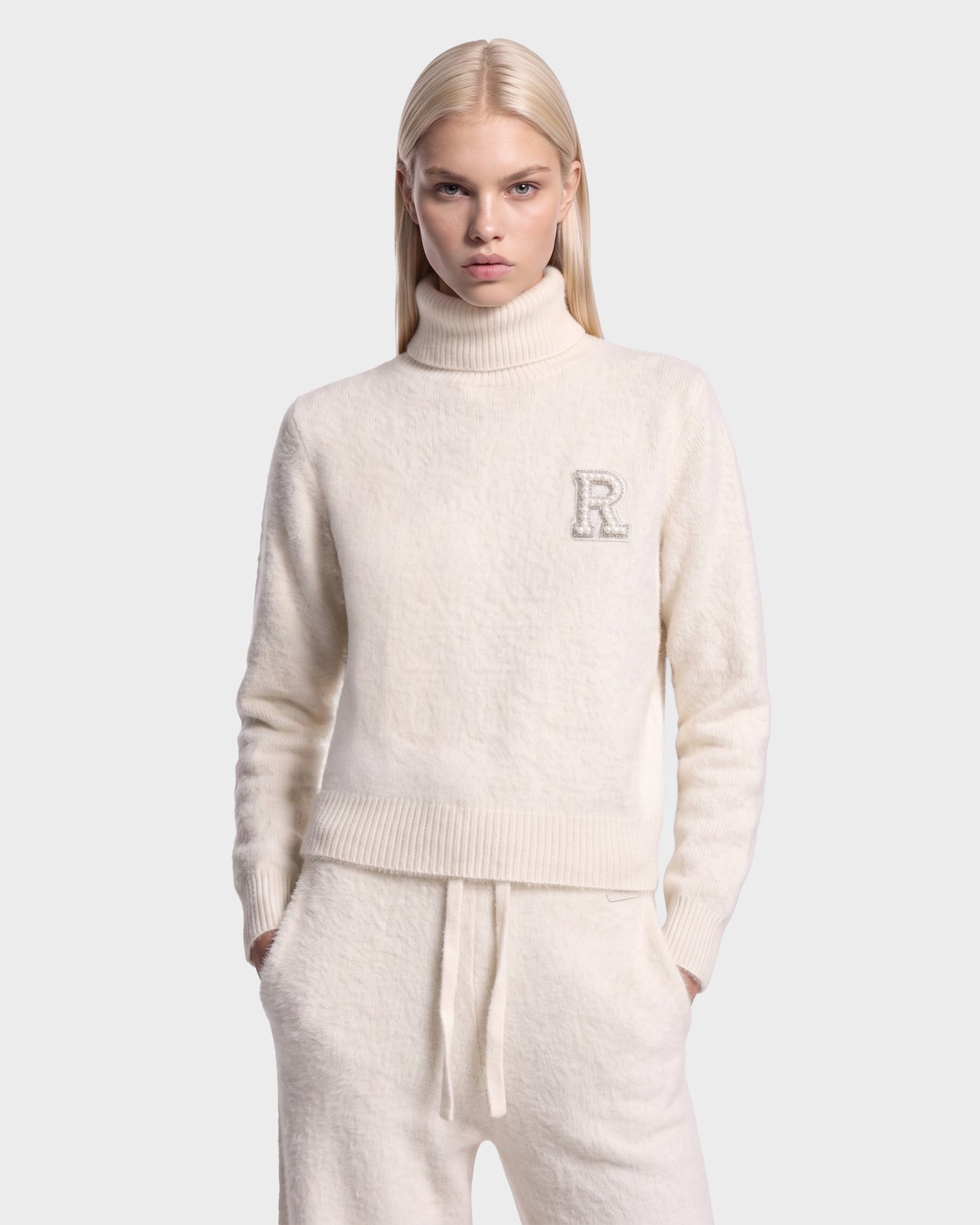 A person with long blonde hair wears the ALISYA - 8526505 Iceland cream-colored turtleneck sweater with an R patch on the left chest and matching drawstring pants by Rudsak, standing against a plain light gray background.