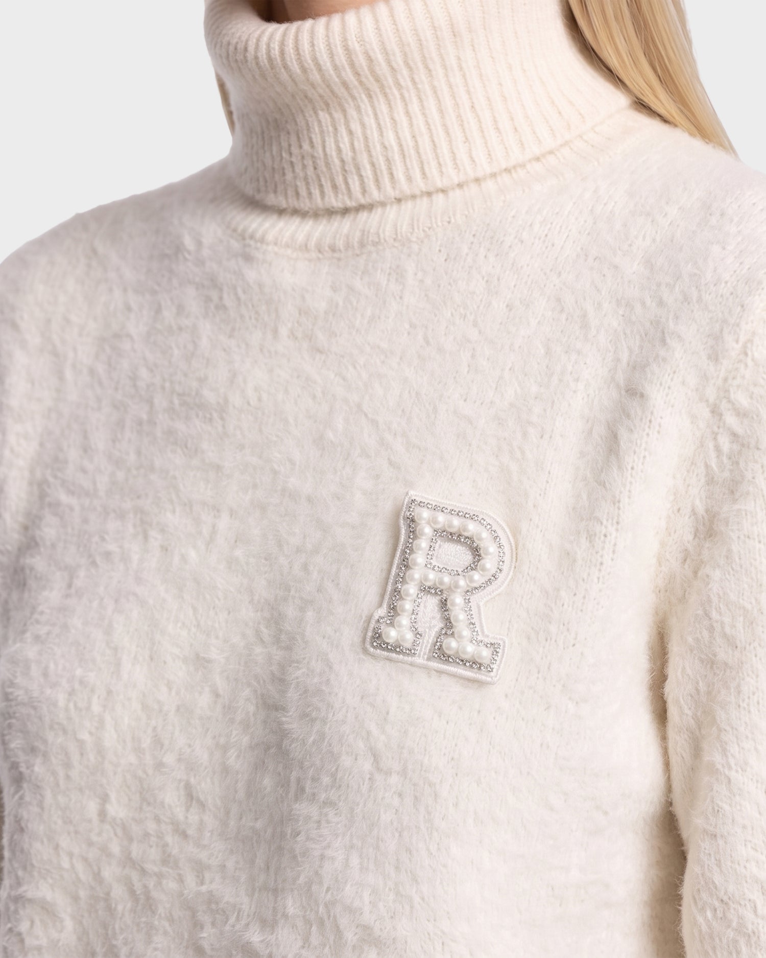 A person wearing the ALISYA - 8526505 Iceland textured white turtleneck sweater by Rudsak, featuring a decorative letter R patch adorned with pearls on the left side of the chest.
