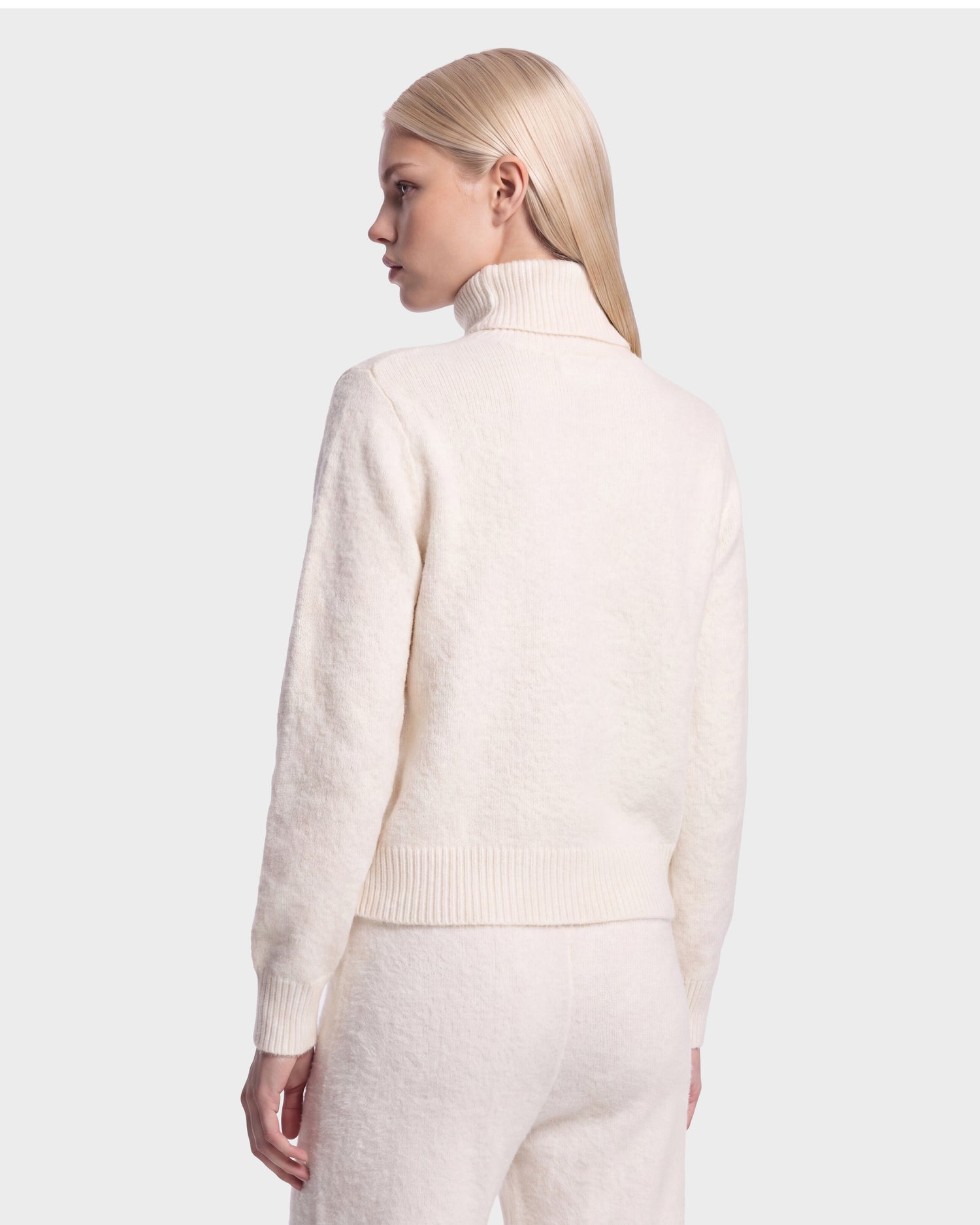 A person with straight, light blond hair wears the Rudsak ALISYA - 8526505 Iceland cream-colored turtleneck sweater and matching pants, standing against a plain light gray background, facing away and looking slightly to the side.