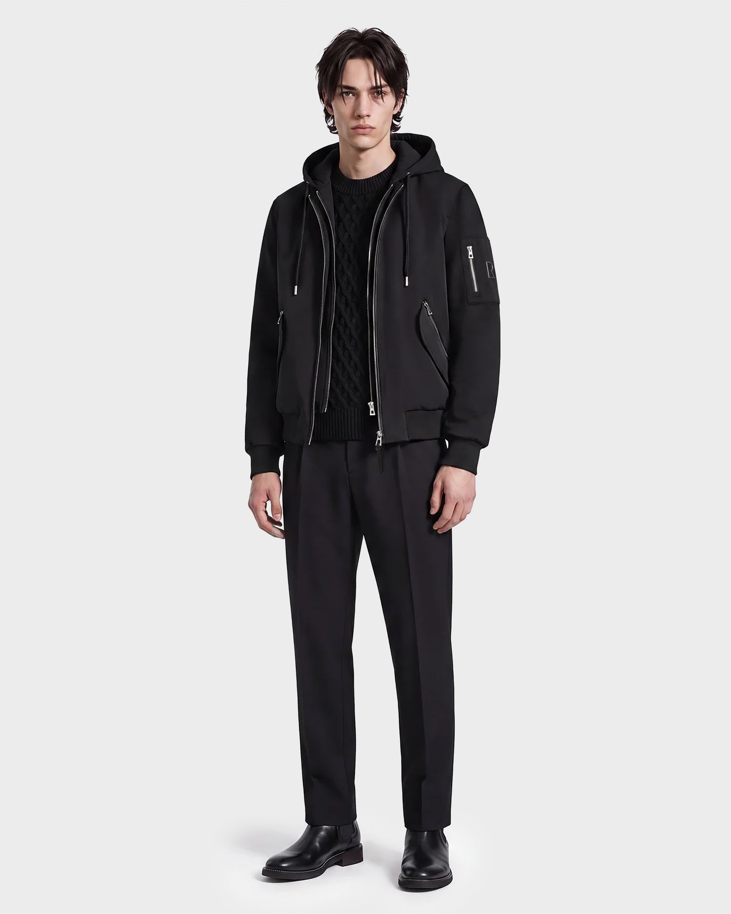 A man stands against a plain background wearing the Rudsak ANDRE - 6124020 Black zip-up hoodie with a removable hooded bib, paired with black pants and shoes, his expression neutral and posture relaxed.