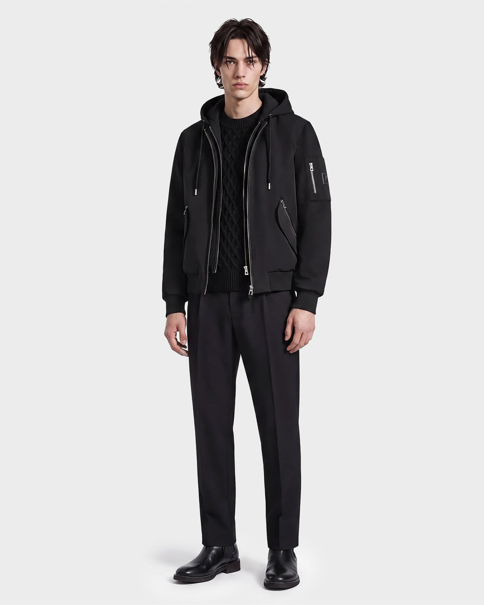 A man stands against a plain background wearing the Rudsak ANDRE - 6124020 Black zip-up hoodie with a removable hooded bib, paired with black pants and shoes, his expression neutral and posture relaxed.