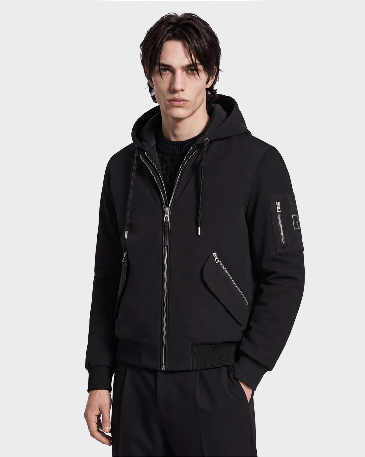 A young man with tousled dark hair models the Rudsak ANDRE - 6124020 Black men’s lightweight bomber jacket, distinguished by zipper accents and a removable hooded bib, paired with matching black pants against a plain white background for a sleek, modern look.
