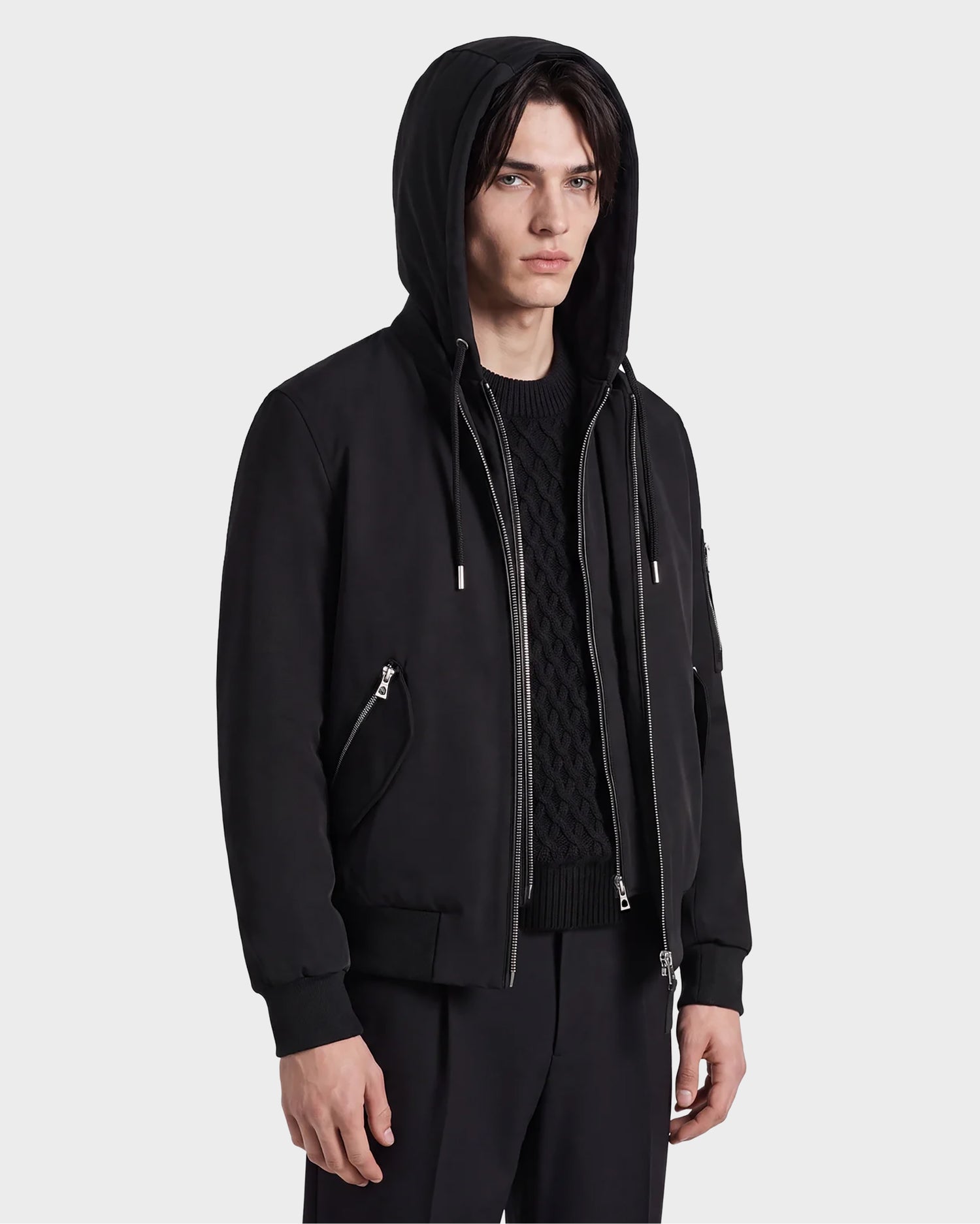 A young man with straight dark hair models the ANDRE - 6124020 Black zip-up hoodie by Rudsak, styled over a black textured sweater and black pants, standing against a plain light background.