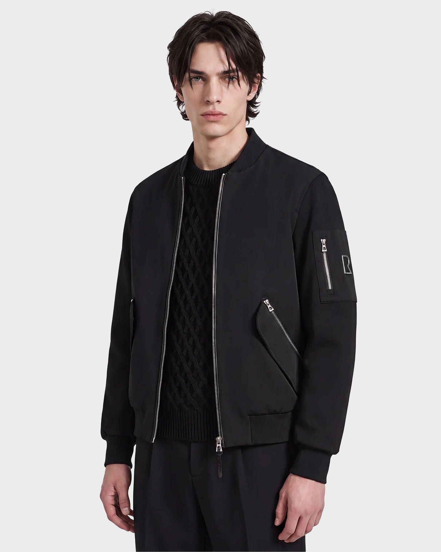 A young man with medium-length brown hair wears the Rudsak ANDRE - 6124020 Black men's bomber jacket over a textured black sweater, standing against a plain white background.