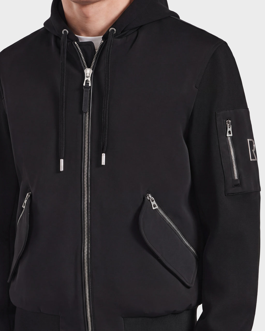 A person wearing the Rudsak ANDRE - 6124020 Black men's lightweight bomber jacket, featuring silver zippers on the front, pockets, and sleeve. The jacket includes a patch on the left upper arm, visible drawstrings on the hood, and a removable hooded bib. The person's face is not shown.