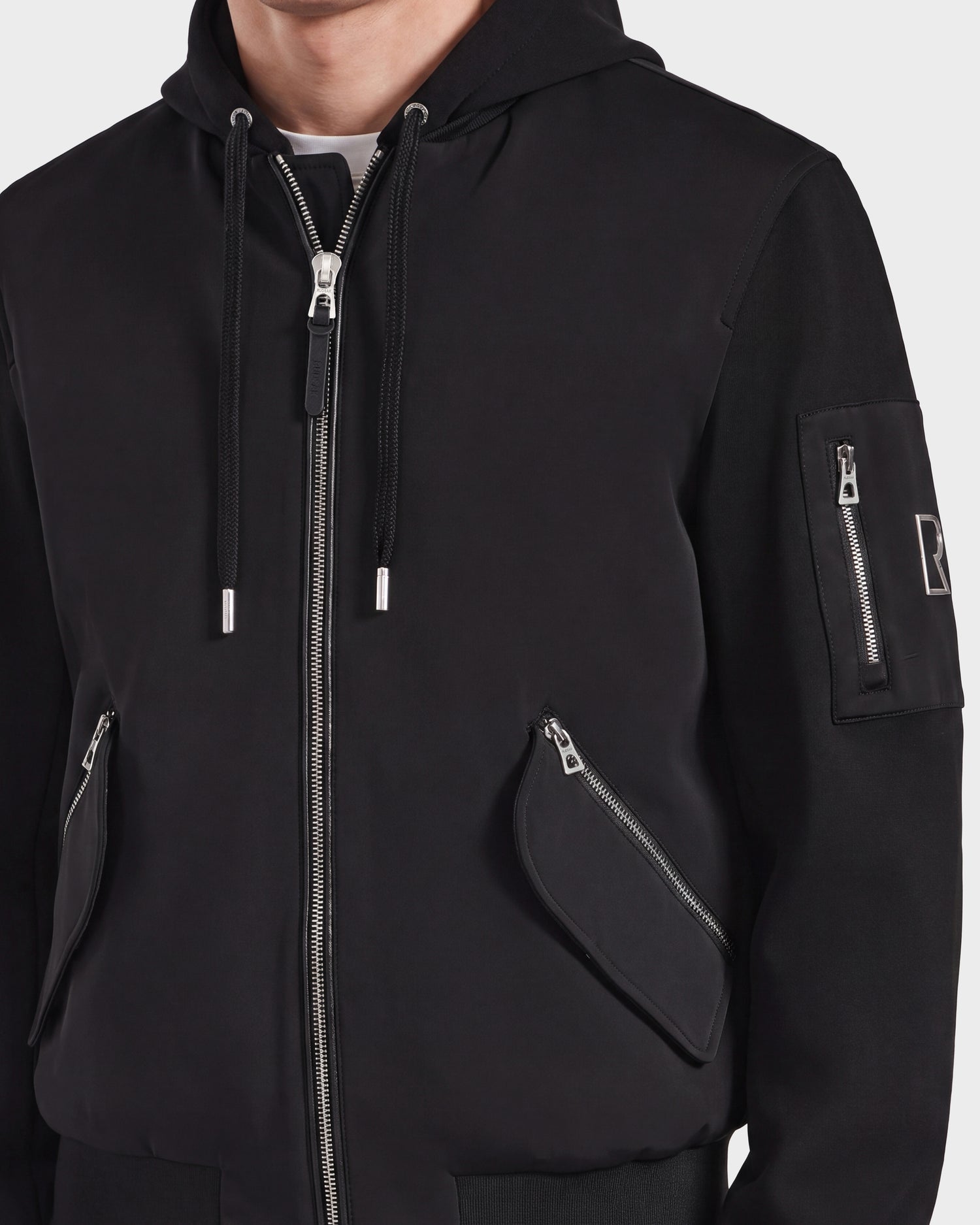 A person wearing the Rudsak ANDRE - 6124020 Black men's lightweight bomber jacket, featuring silver zippers on the front, pockets, and sleeve. The jacket includes a patch on the left upper arm, visible drawstrings on the hood, and a removable hooded bib. The person's face is not shown.