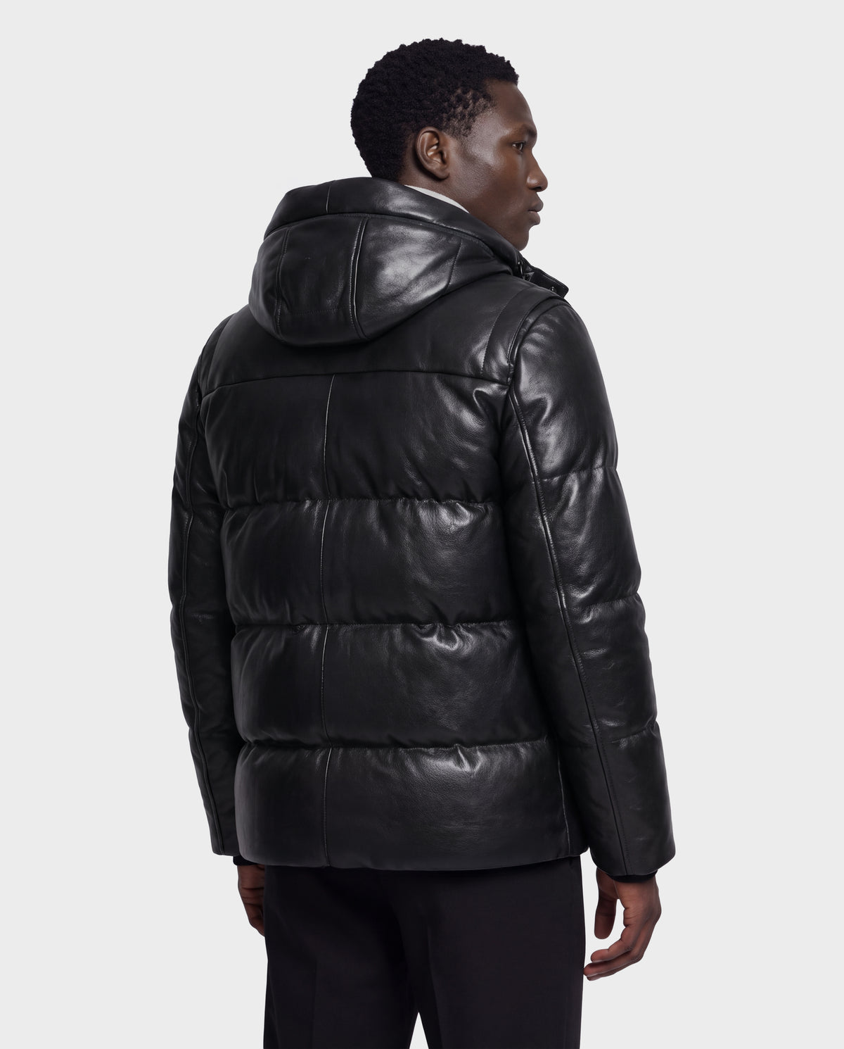 A person stands facing away, wearing the APOLLO X - 6125696 Black men’s leather down jacket from Rudsak. The jacket features a shiny black, quilted design with a hood and appears thick and insulated, suggesting it contains RDS-certified down for enhanced warmth. The background is plain white.