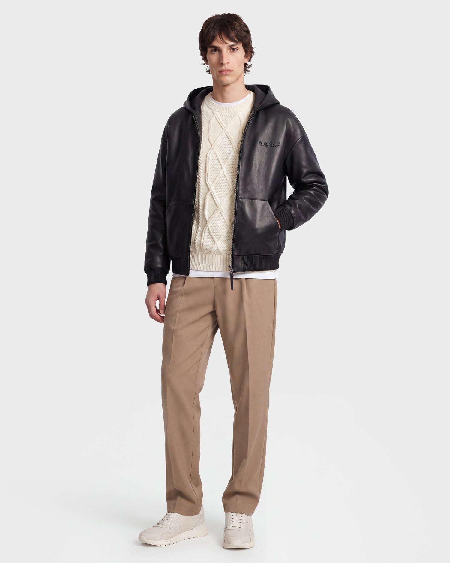 A person stands against a plain white background, wearing the Rudsak ARCHIE - 6126500 Black leather jacket over a white cable-knit sweater, paired with tan pants and white sneakers.