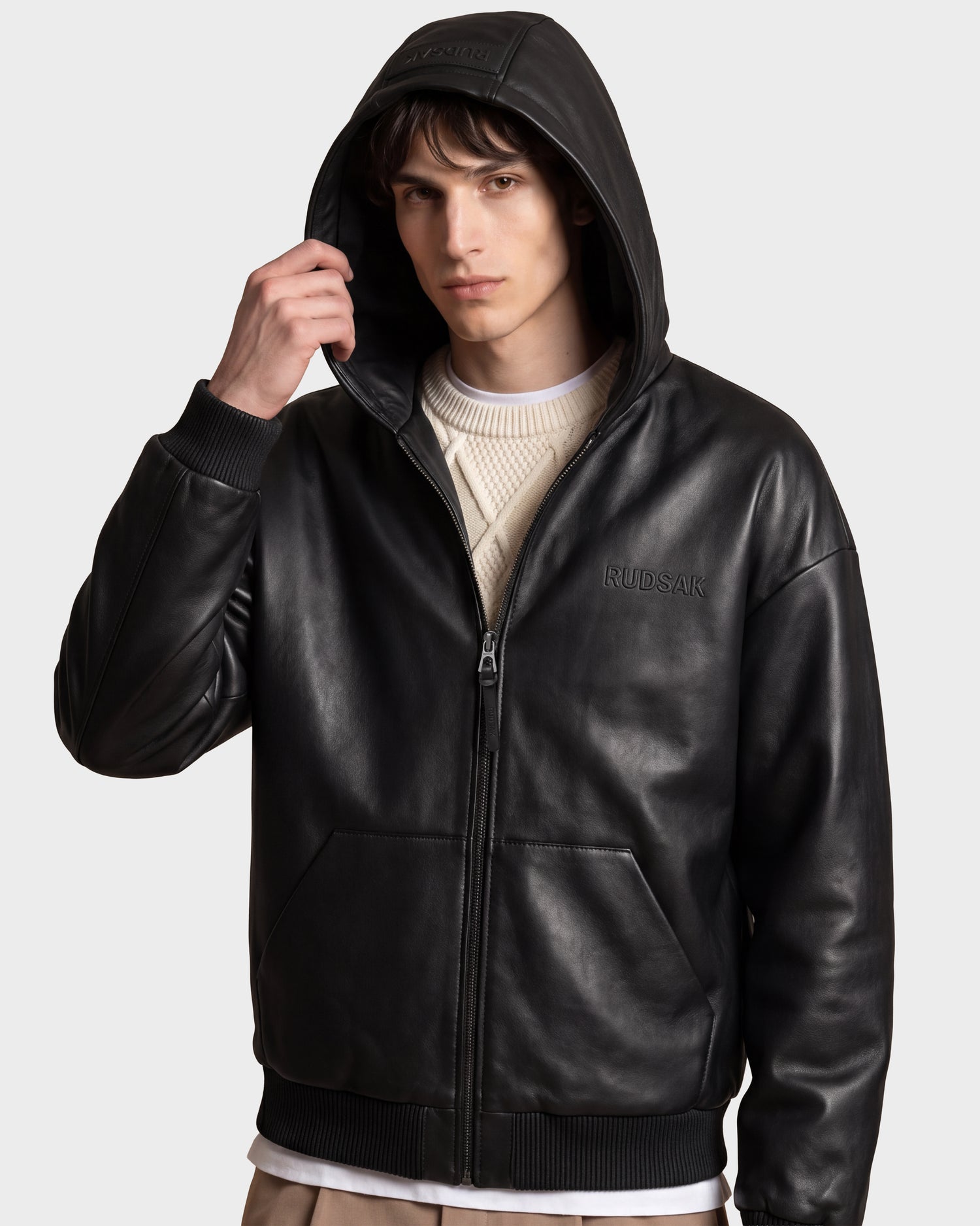 A young man stands with one hand adjusting the hood of the Rudsak ARCHIE - 6126500 Black leather zip-up hoodie, worn over a cream knit sweater and paired with light brown pants.