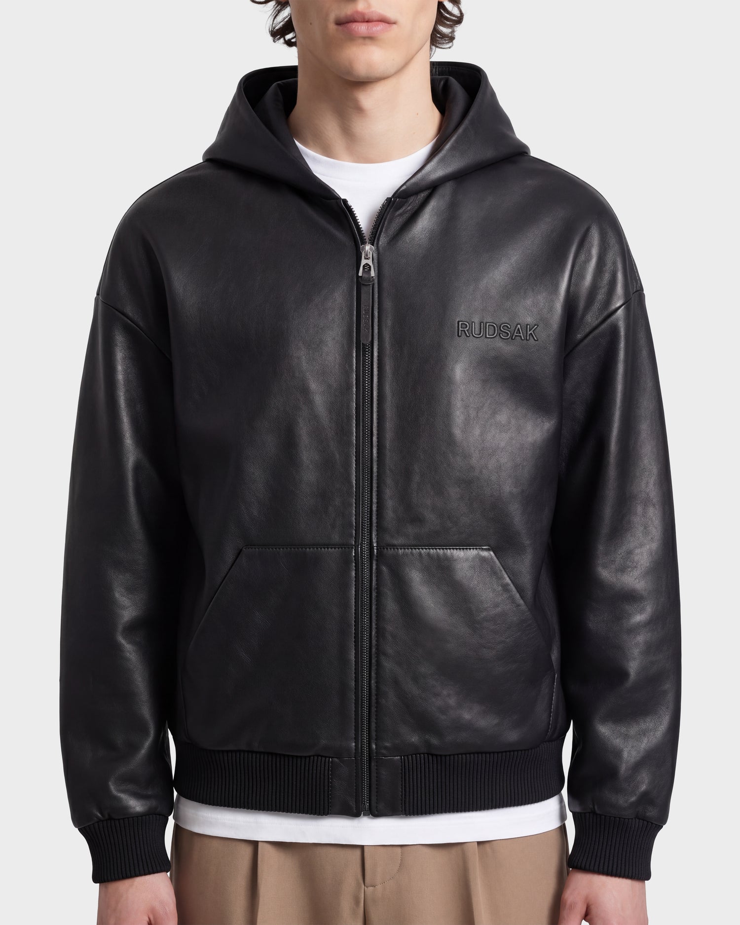 A person wears the Rudsak ARCHIE - 6126500 Black leather zip-up hoodie, featuring front pockets and an embroidered RUDSAK logo on the chest, layered over a white shirt and tan pants, set against a plain light background.