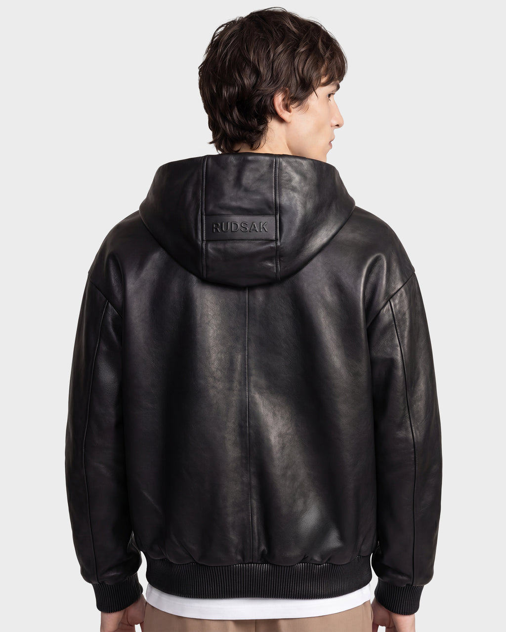 A person with short brown hair is seen from the back wearing the ARCHIE - 6126500 Black hooded leather jacket by Rudsak, featuring "RUDSAK" embossed on the hood, standing against a plain light background.