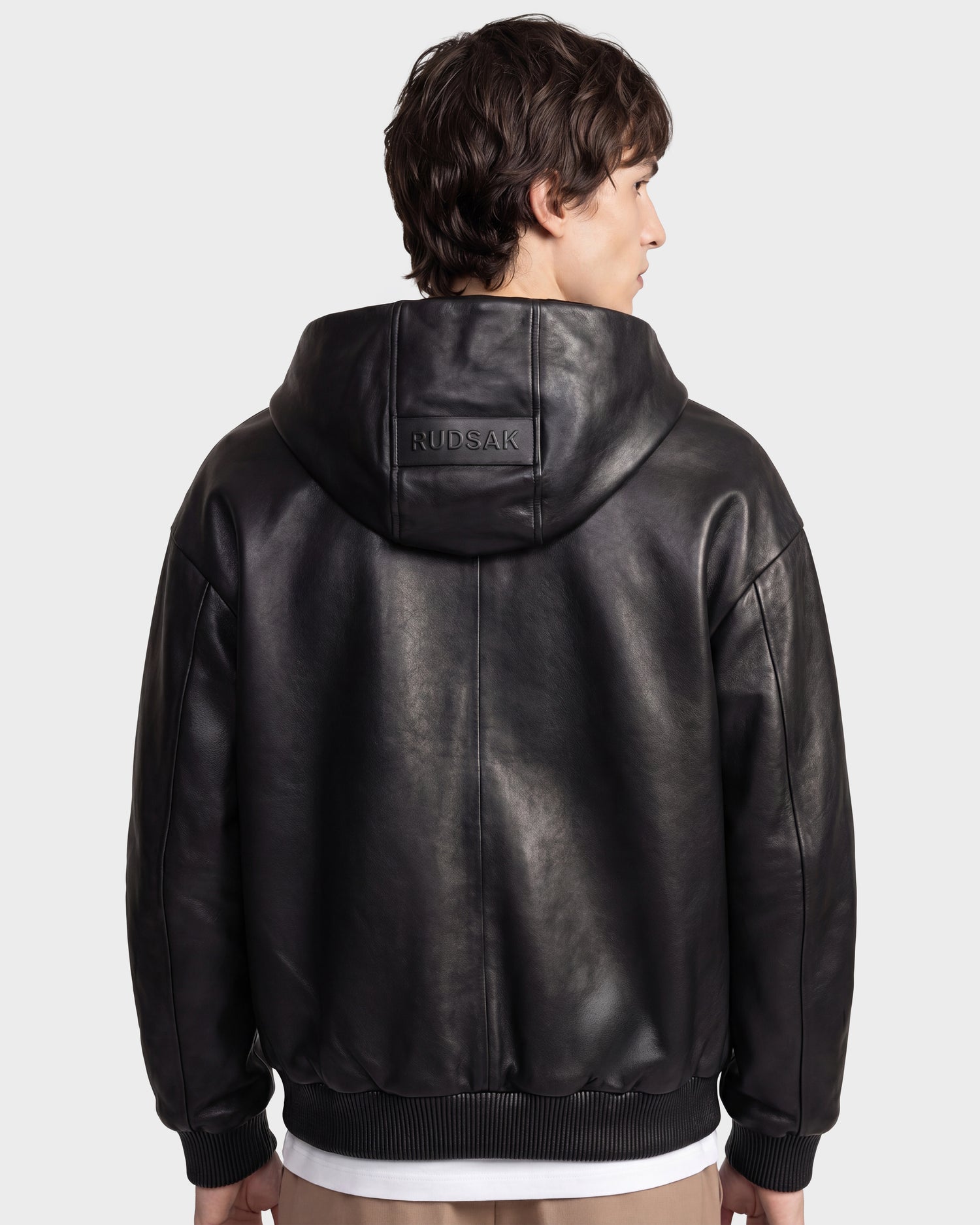 A person with short brown hair is seen from the back wearing the ARCHIE - 6126500 Black hooded leather jacket by Rudsak, featuring "RUDSAK" embossed on the hood, standing against a plain light background.