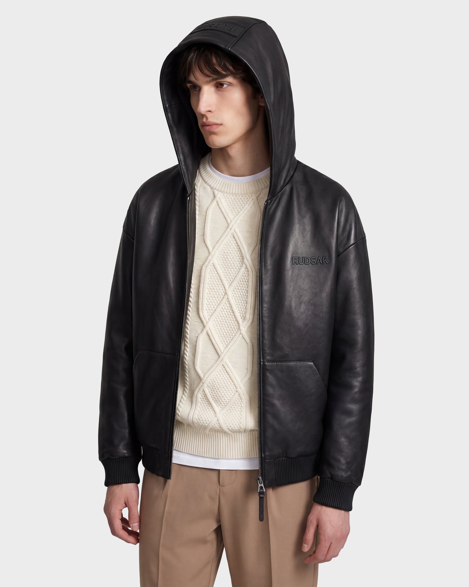 A person wearing the Rudsak ARCHIE - 6126500 Black leather hooded jacket over a white cable-knit sweater and beige pants stands against a plain light background, looking to the side.