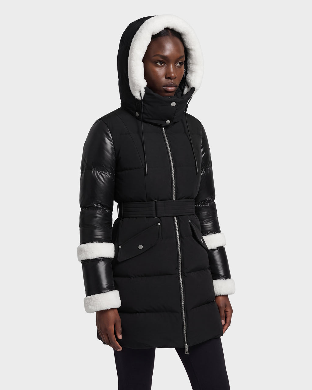 A woman wearing the Rudsak AUDREY L - 8124585 Black/Iceland puffer jacket, featuring recycled Stormshell fabric, a belted waist, faux leather sleeves, and white faux fur trim on the hood and cuffs, stands against a plain light background.