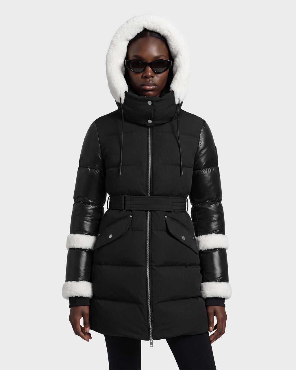 A person stands straight against a light gray background wearing the Rudsak AUDREY L - 8124585 Black/Iceland, a knee-length down puffer jacket featuring a white fur-lined hood, leather sleeve trim, black belt, and black sunglasses. The coat is crafted from recycled Stormshell fabric and insulated with RDS certified down for warmth and sustainability.