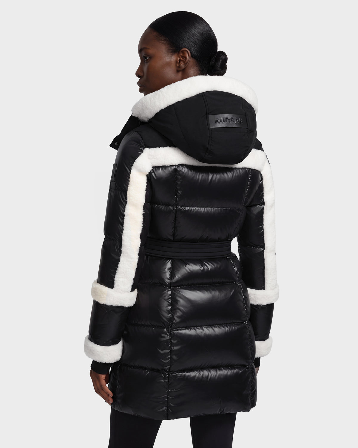 A woman stands facing away, showcasing the back of the Rudsak AUDREY L - 8124585 Black/Iceland knee-length down puffer jacket, crafted from recycled Stormshell fabric with white trim and a hood.