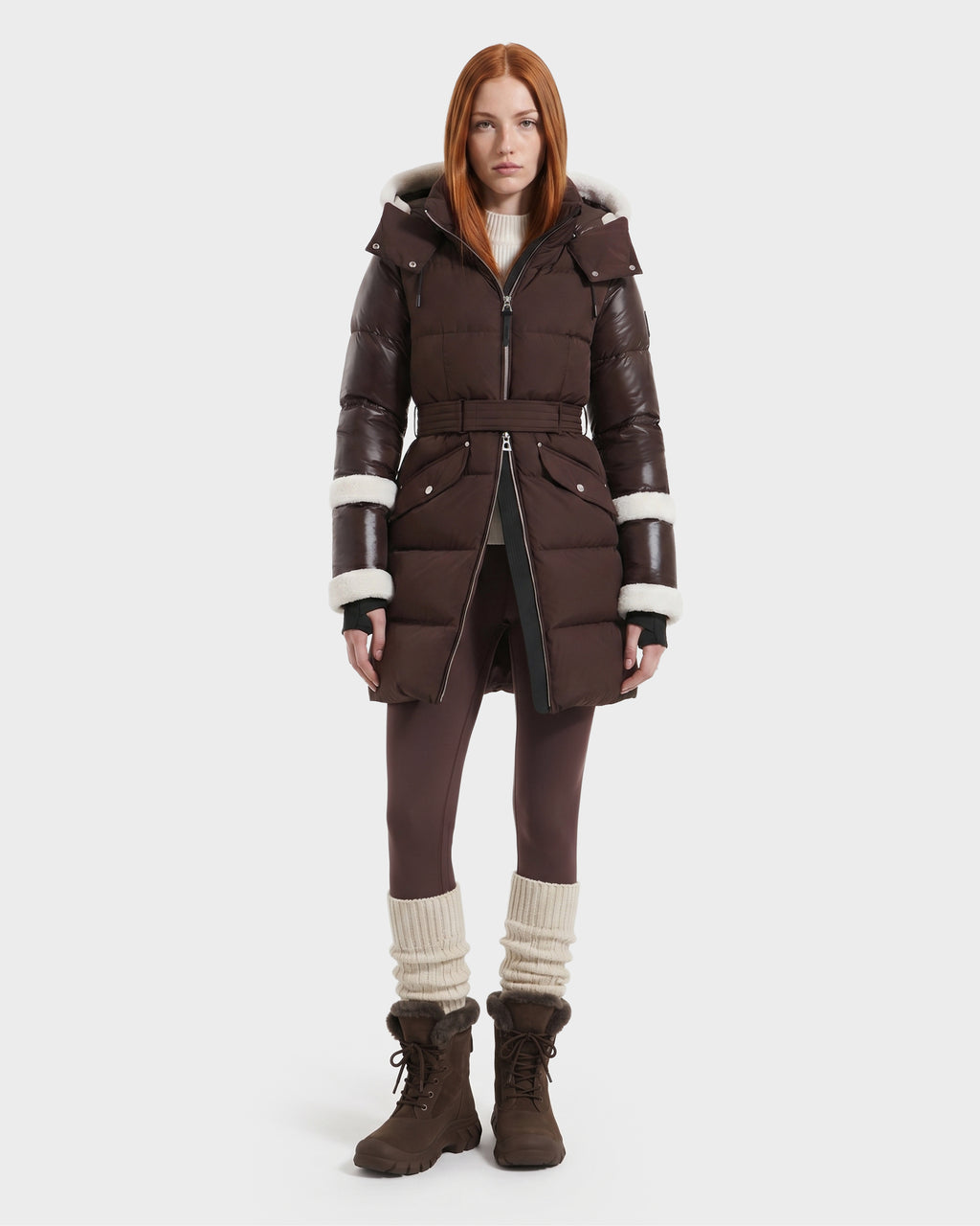 A woman with straight red hair models the Rudsak AUDREY L - 8124585 Espresso, a knee-length down puffer jacket crafted from recycled Stormshell fabric and filled with RDS-certified down. The jacket features rich brown tones and a white fur trim. She pairs it with brown leggings, tall slouchy socks, and brown boots while standing against a plain white background.