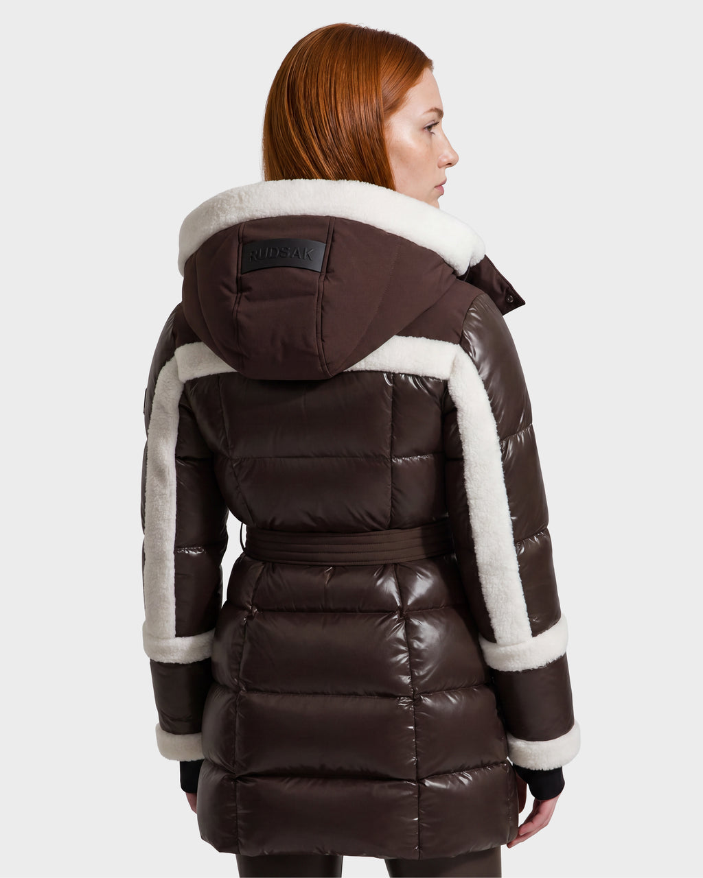 A woman with straight red hair models the Rudsak AUDREY L - 8124585 Espresso, a knee-length down puffer jacket crafted from recycled Stormshell fabric. Featuring a hood, white shearling trim, and a belted waist, the dark brown coat is shown from the back against a plain light gray background.