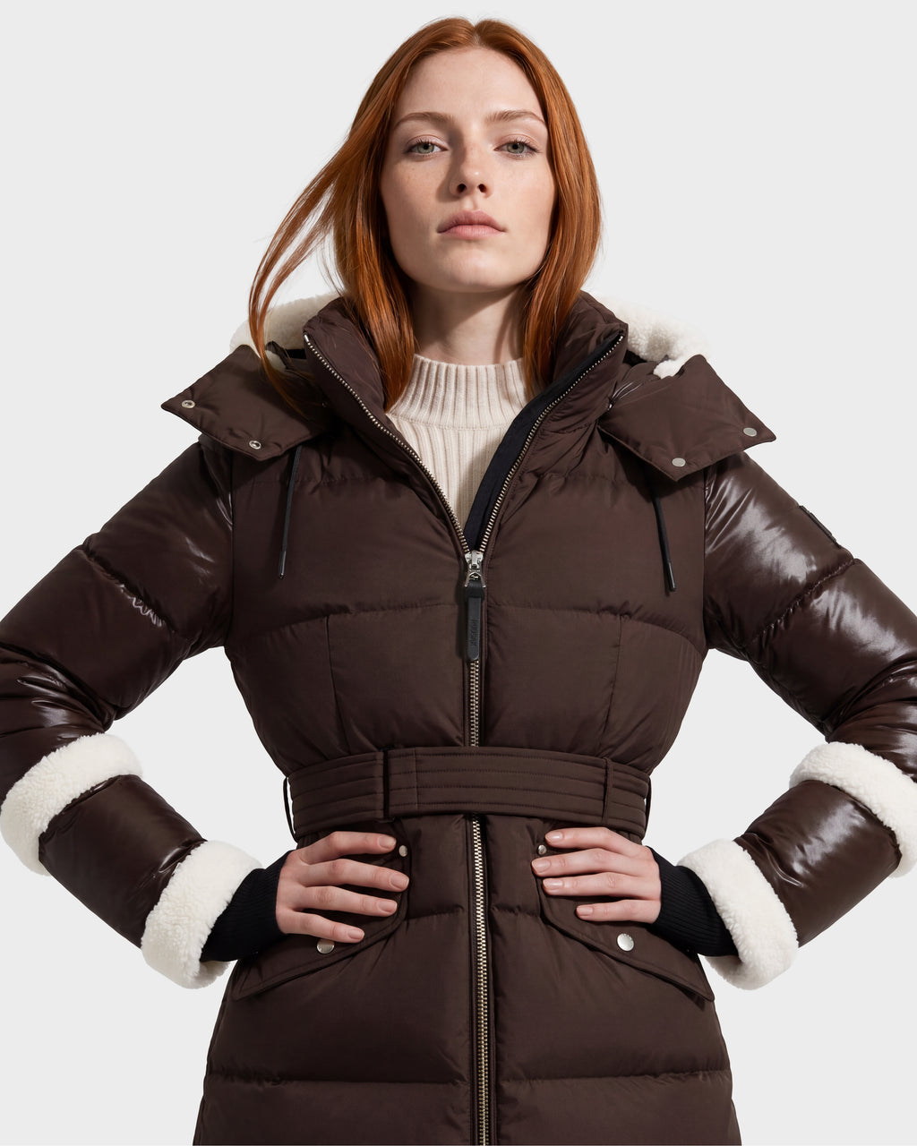 A woman with long red hair models the Rudsak AUDREY L - 8124585 Espresso knee-length down puffer jacket, showcasing its belted waist and white fuzzy trim on the hood and cuffs. Crafted from recycled Stormshell fabric and filled with RDS certified down, this brown coat is layered over a beige knit turtleneck sweater as she poses against a plain light background.