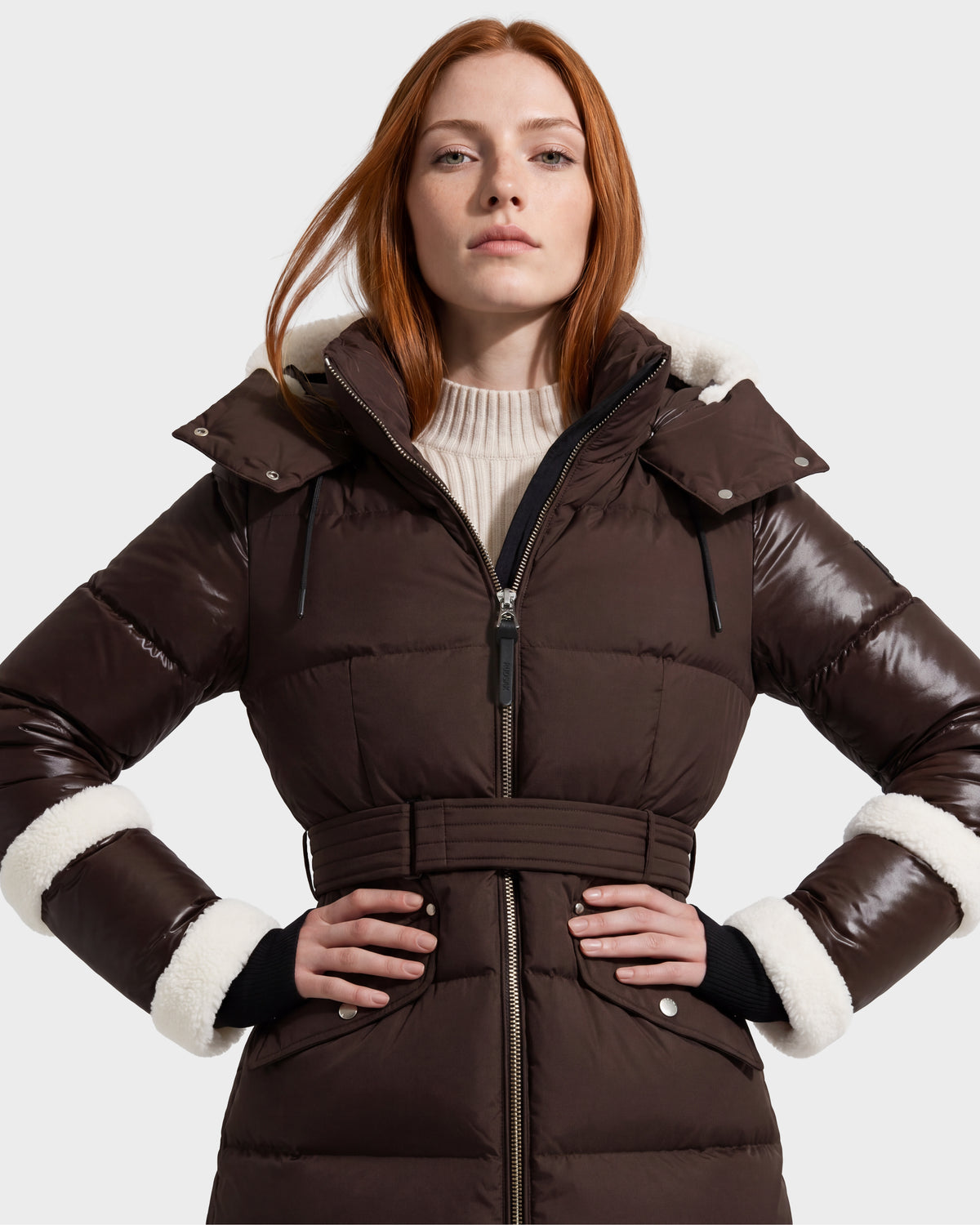 A woman with long red hair models the Rudsak AUDREY L - 8124585 Espresso knee-length down puffer jacket, showcasing its belted waist and white fuzzy trim on the hood and cuffs. Crafted from recycled Stormshell fabric and filled with RDS certified down, this brown coat is layered over a beige knit turtleneck sweater as she poses against a plain light background.