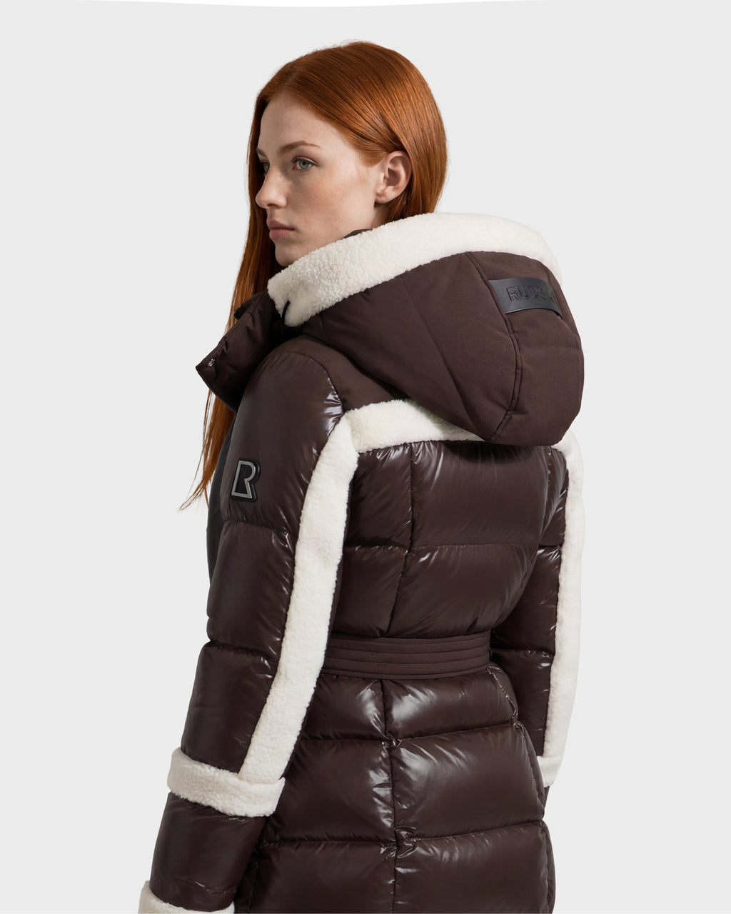 A woman with long red hair looks to the side against a plain light gray background, wearing the Rudsak AUDREY L - 8124585 Espresso—a shiny brown knee-length down puffer jacket crafted from recycled Stormshell fabric and finished with white shearling trim.