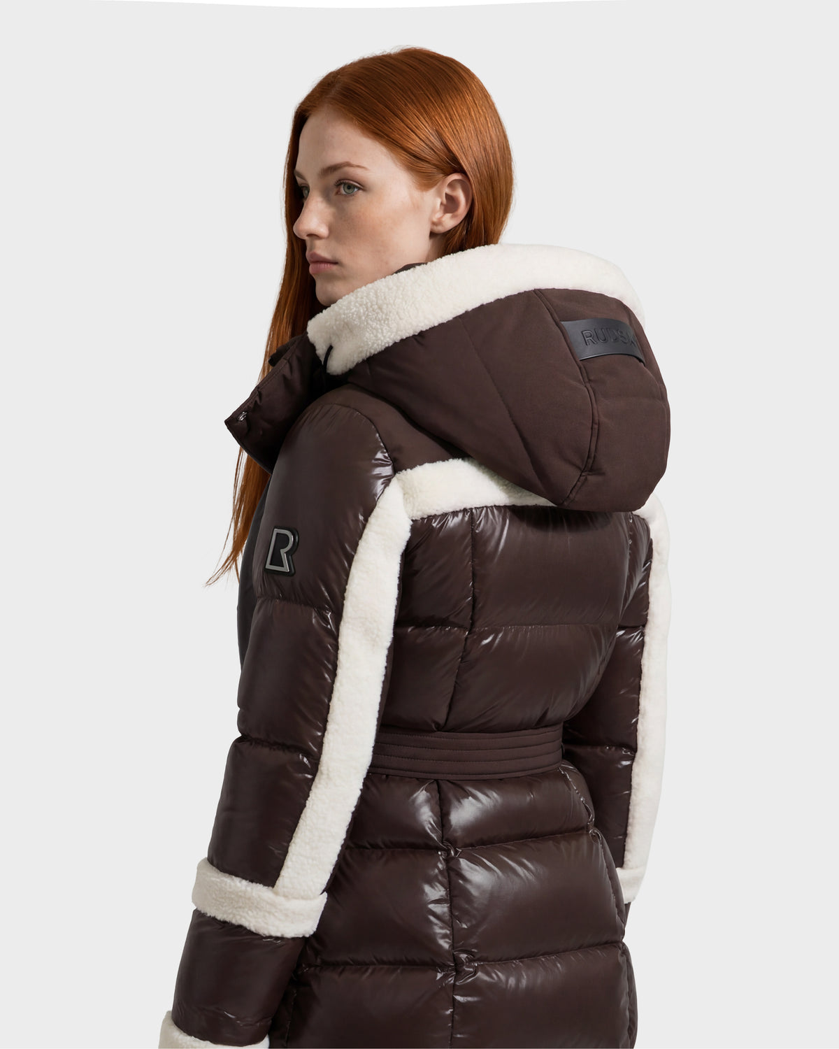 A woman with long red hair looks to the side against a plain light gray background, wearing the Rudsak AUDREY L - 8124585 Espresso—a shiny brown knee-length down puffer jacket crafted from recycled Stormshell fabric and finished with white shearling trim.