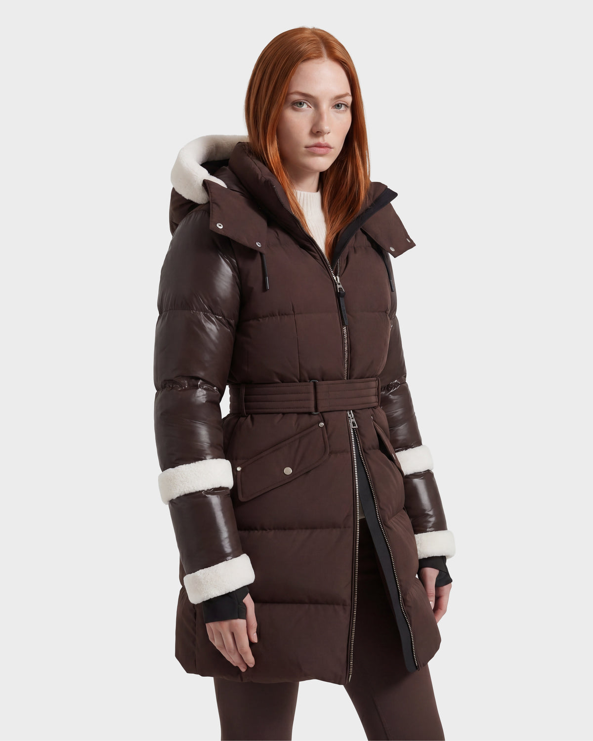 A woman with straight red hair models the Rudsak AUDREY L - 8124585 Espresso, a knee-length down puffer jacket made from recycled Stormshell fabric. The jacket features a long, brown, belted silhouette and is accented with white faux fur trim on the hood and cuffs. She stands against a plain light background.