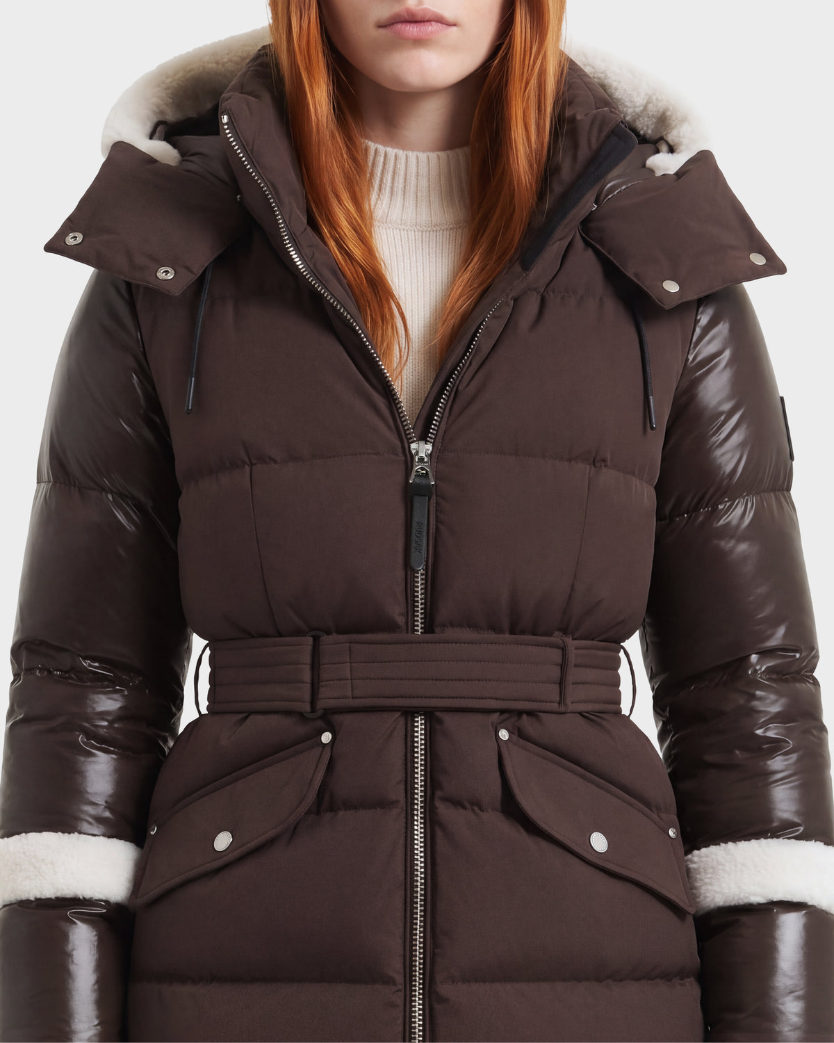 A woman with long red hair wears the Rudsak AUDREY L - 8124585 puffer jacket in Espresso, a knee-length design crafted from recycled Stormshell fabric. The jacket features shiny sleeves, a hood edged with shearling trim, snap-flap pockets, and a belted waist, layered over a cream turtleneck sweater. Her face is partially out of frame.
