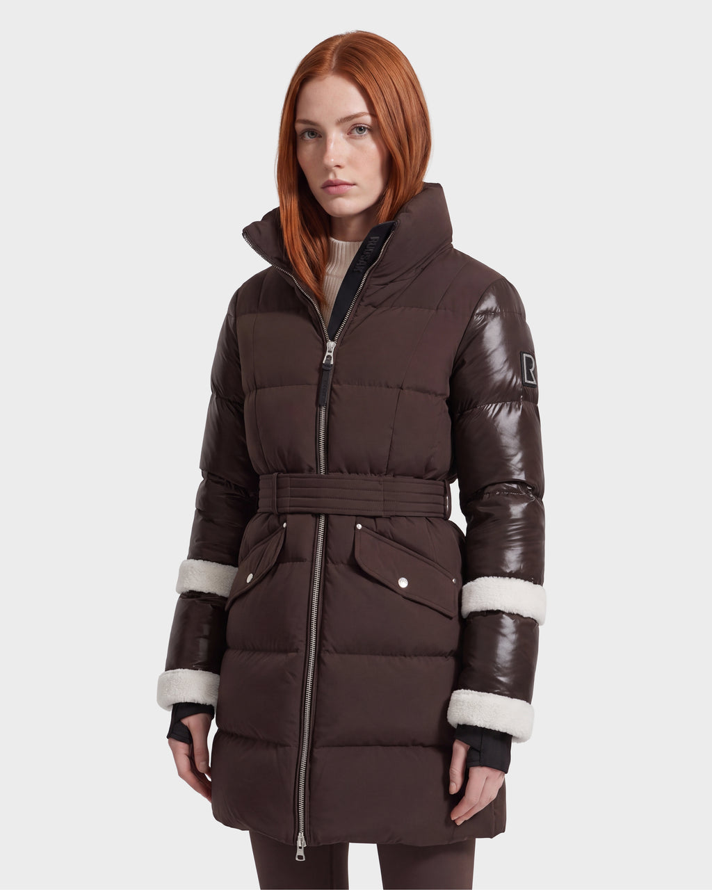 A woman with long red hair models the Rudsak AUDREY L - 8124585 Espresso, a knee-length down puffer jacket featuring a dark brown belted silhouette, glossy sleeves, and white trim detail—crafted from recycled Stormshell fabric—while standing against a plain light background.