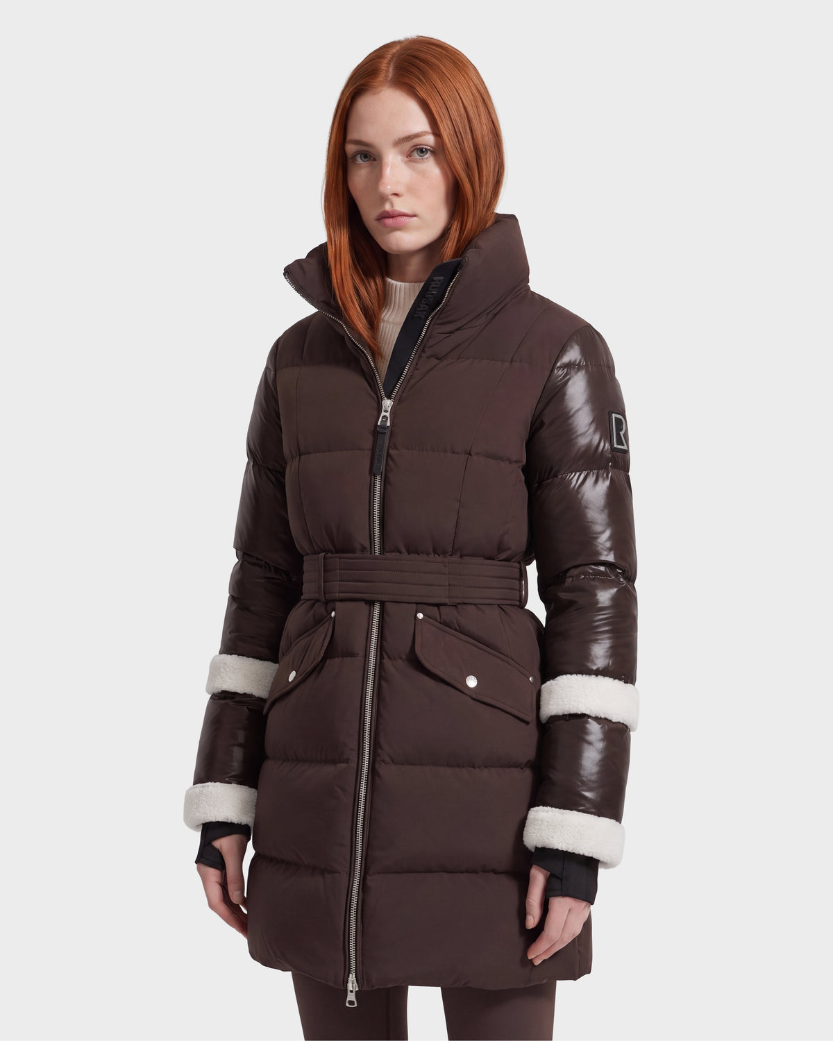 A woman with long red hair models the Rudsak AUDREY L - 8124585 Espresso, a knee-length down puffer jacket featuring a dark brown belted silhouette, glossy sleeves, and white trim detail—crafted from recycled Stormshell fabric—while standing against a plain light background.