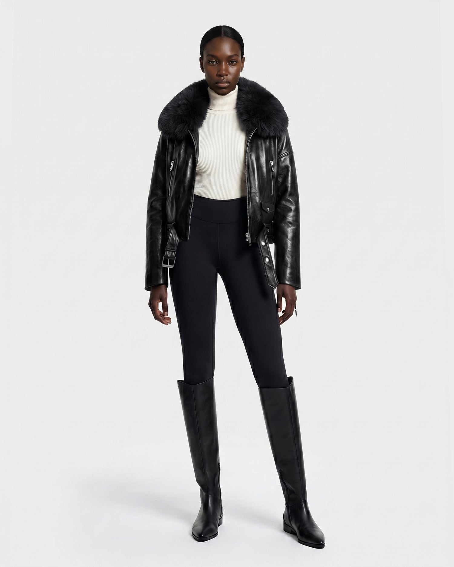 A woman stands against a plain white background wearing the Rudsak AVITA - 8126938 Black leather jacket with a fur collar, paired with a white turtleneck sweater, black fitted pants, and tall black boots.