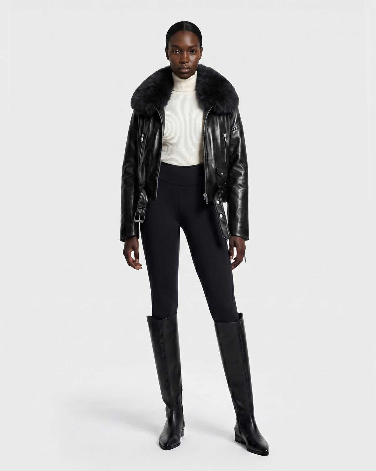 A woman stands against a plain white background wearing the Rudsak AVITA - 8126938 Black leather jacket with a fur collar, paired with a white turtleneck sweater, black fitted pants, and tall black boots.