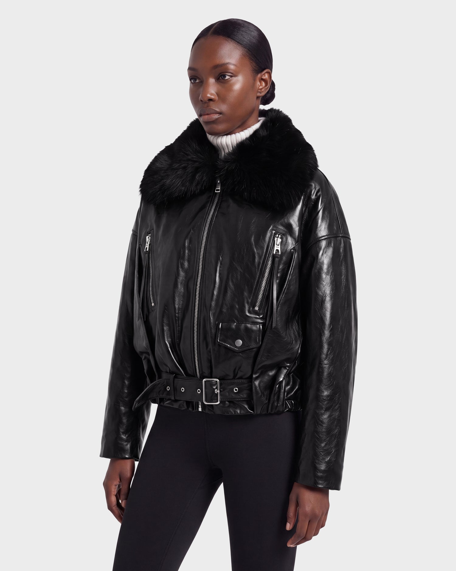 A person wearing the Rudsak AVITA - 8126938 Black leather jacket with a large fur collar, paired with a white turtleneck sweater and black pants, stands against a plain light gray background.