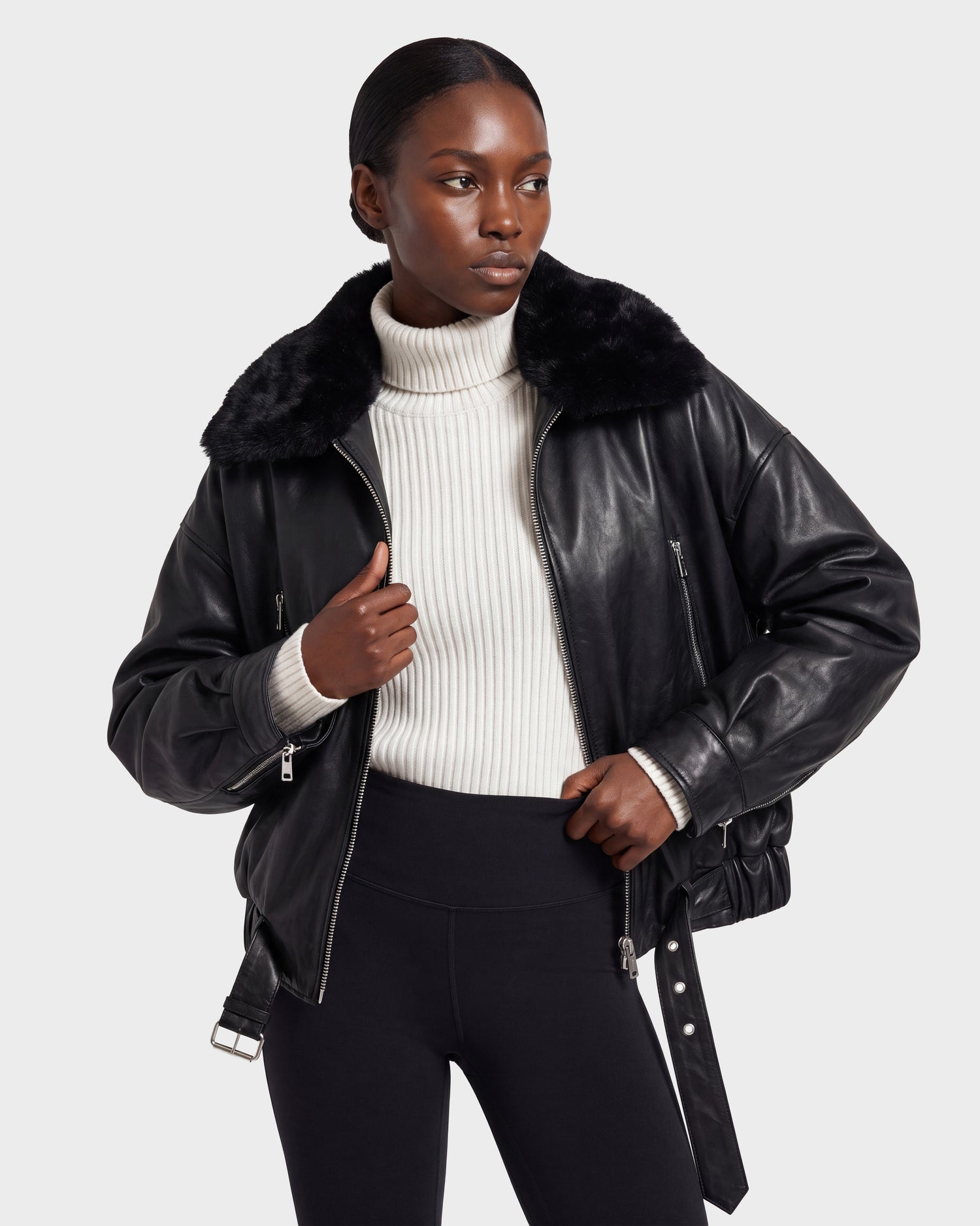 A woman stands against a plain background with one hand on her hip, wearing the Rudsak AVITA - 8126938 Black leather jacket with a fur collar, styled over a white ribbed turtleneck sweater and black pants.