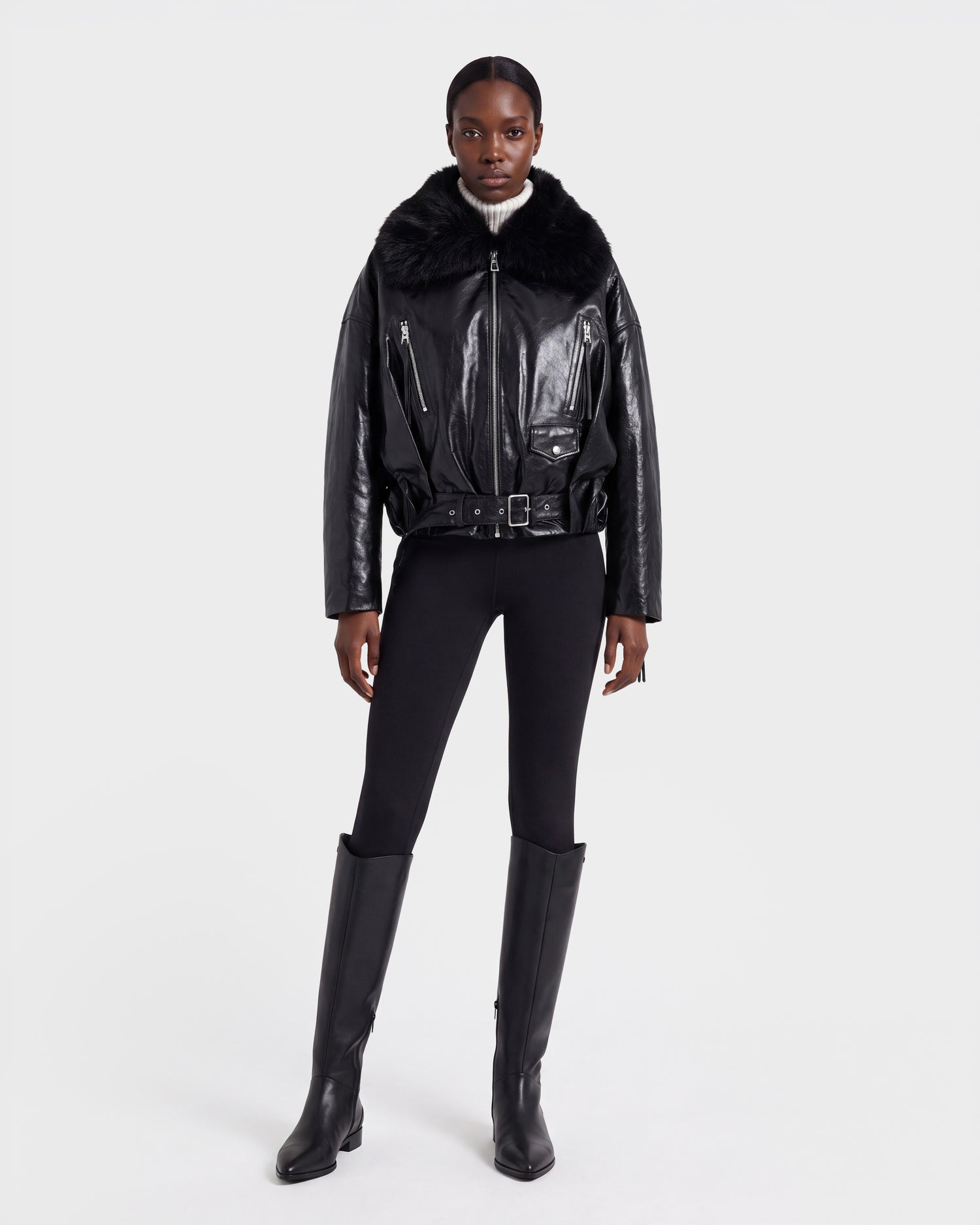 A woman stands against a plain white background wearing the Rudsak AVITA - 8126938 Black leather jacket with a fur collar, paired with black leggings and knee-high black boots.