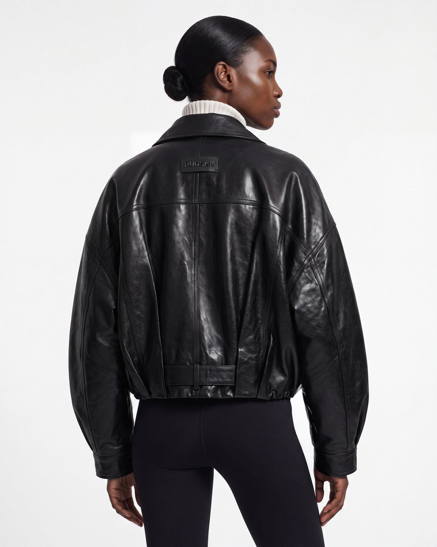 A person with sleek, dark hair styled in a low bun faces away, wearing the Rudsak AVITA - 8126938 Black leather jacket and black leggings. The jacket features a patch on the upper back and reveals a ribbed turtleneck underneath, set against a plain white background.