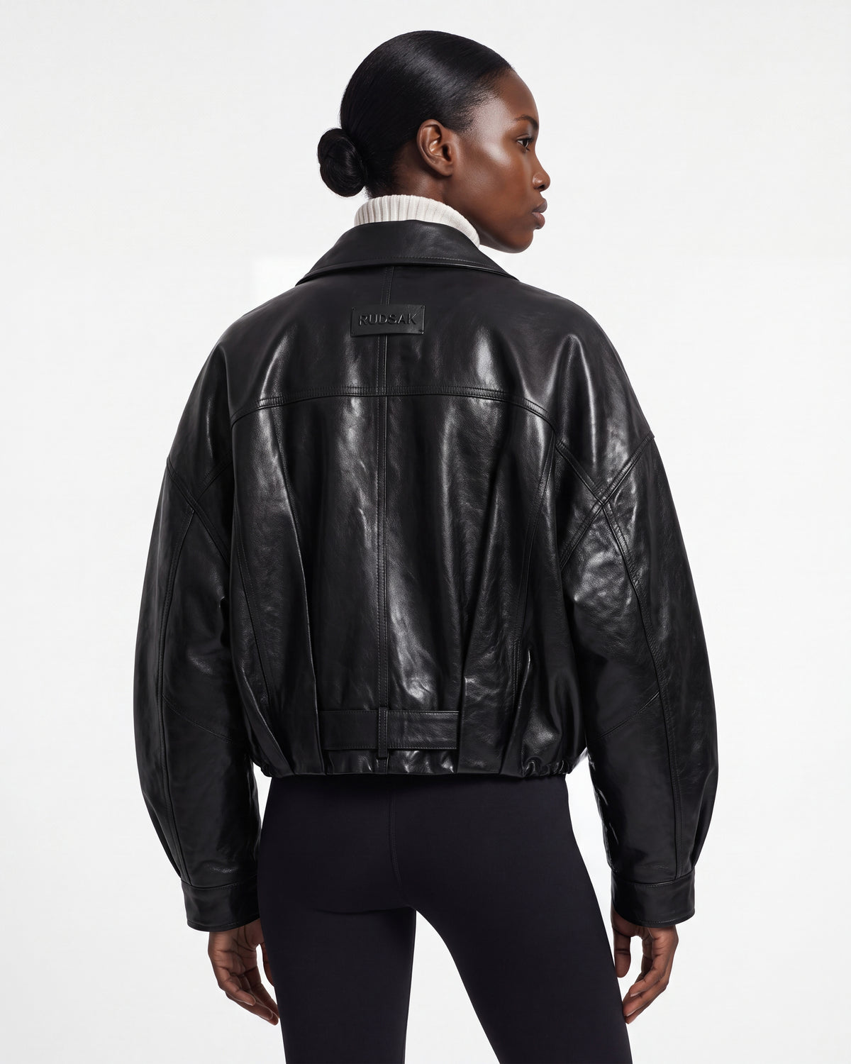 A person with sleek, dark hair styled in a low bun faces away, wearing the Rudsak AVITA - 8126938 Black leather jacket and black leggings. The jacket features a patch on the upper back and reveals a ribbed turtleneck underneath, set against a plain white background.