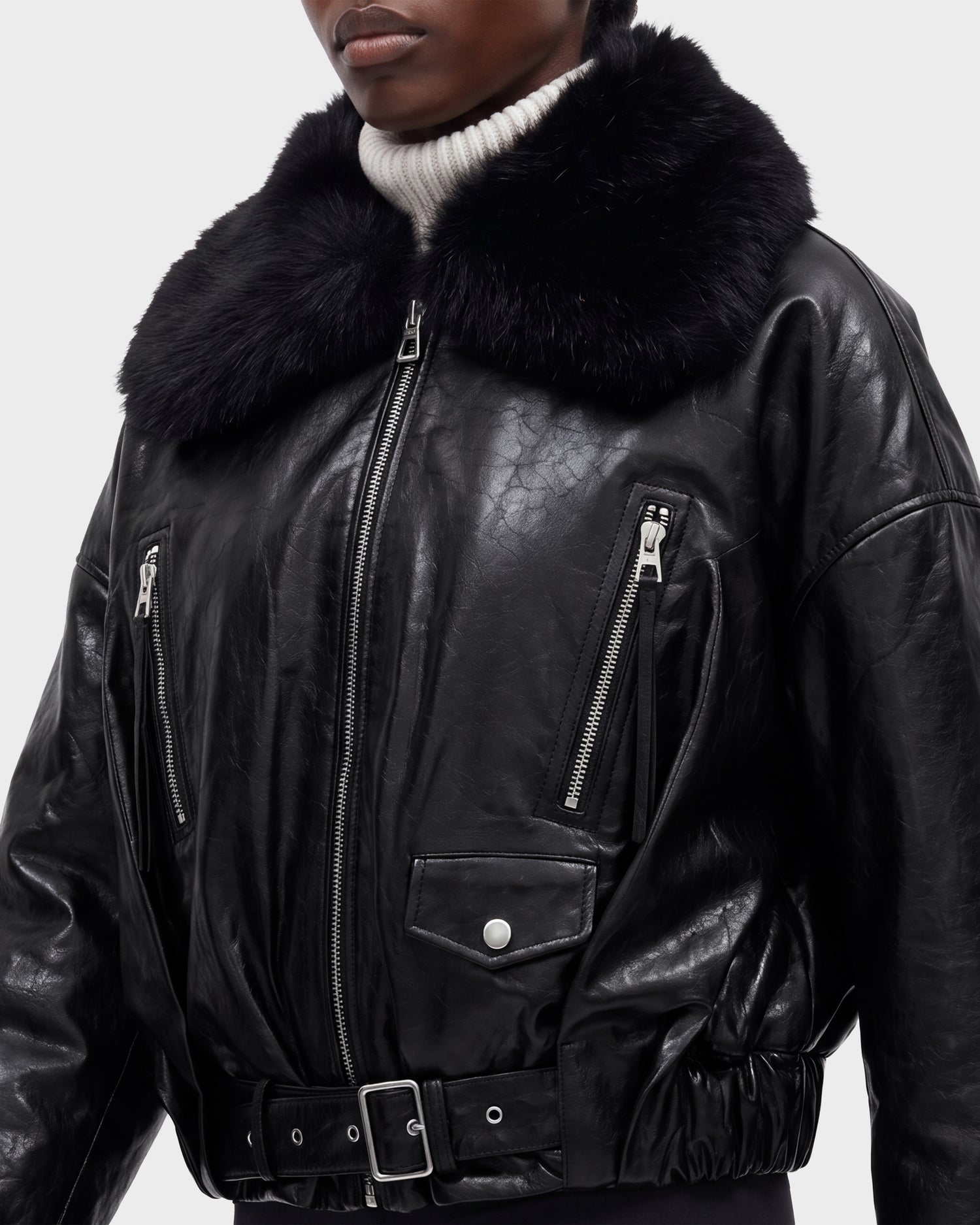 A person wearing the Rudsak AVITA - 8126938 Black leather jacket, which features a wide, fuzzy black collar, zippered pockets, and a belted waist, over a white ribbed turtleneck sweater.