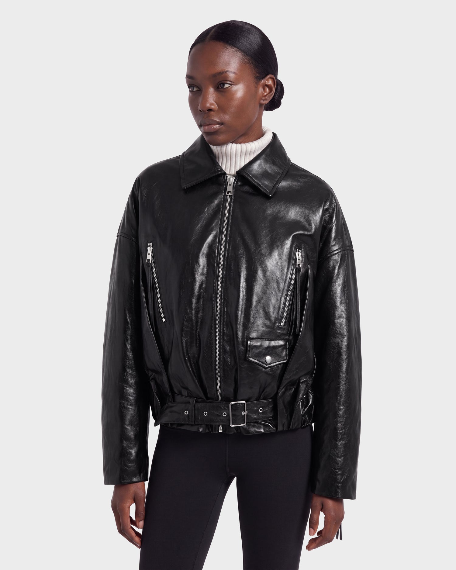 A person wearing the Rudsak AVITA - 8126938 Black leather jacket, featuring zippered pockets and a waist belt, paired with black pants and a white turtleneck, stands against a plain light gray background.
